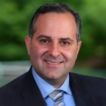 A professional headshot of a man in a dark suit and light blue shirt, smiling, with a green blurred outdoor background.