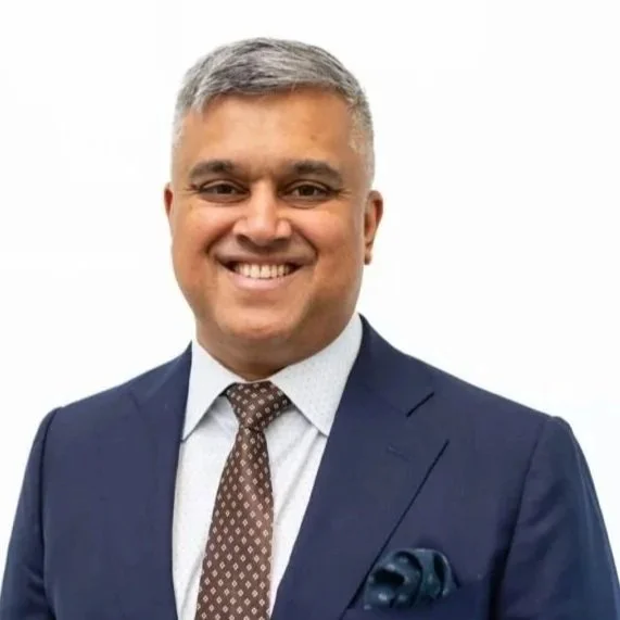 A man in a navy suit, white shirt, and patterned tie smiling against a white background.