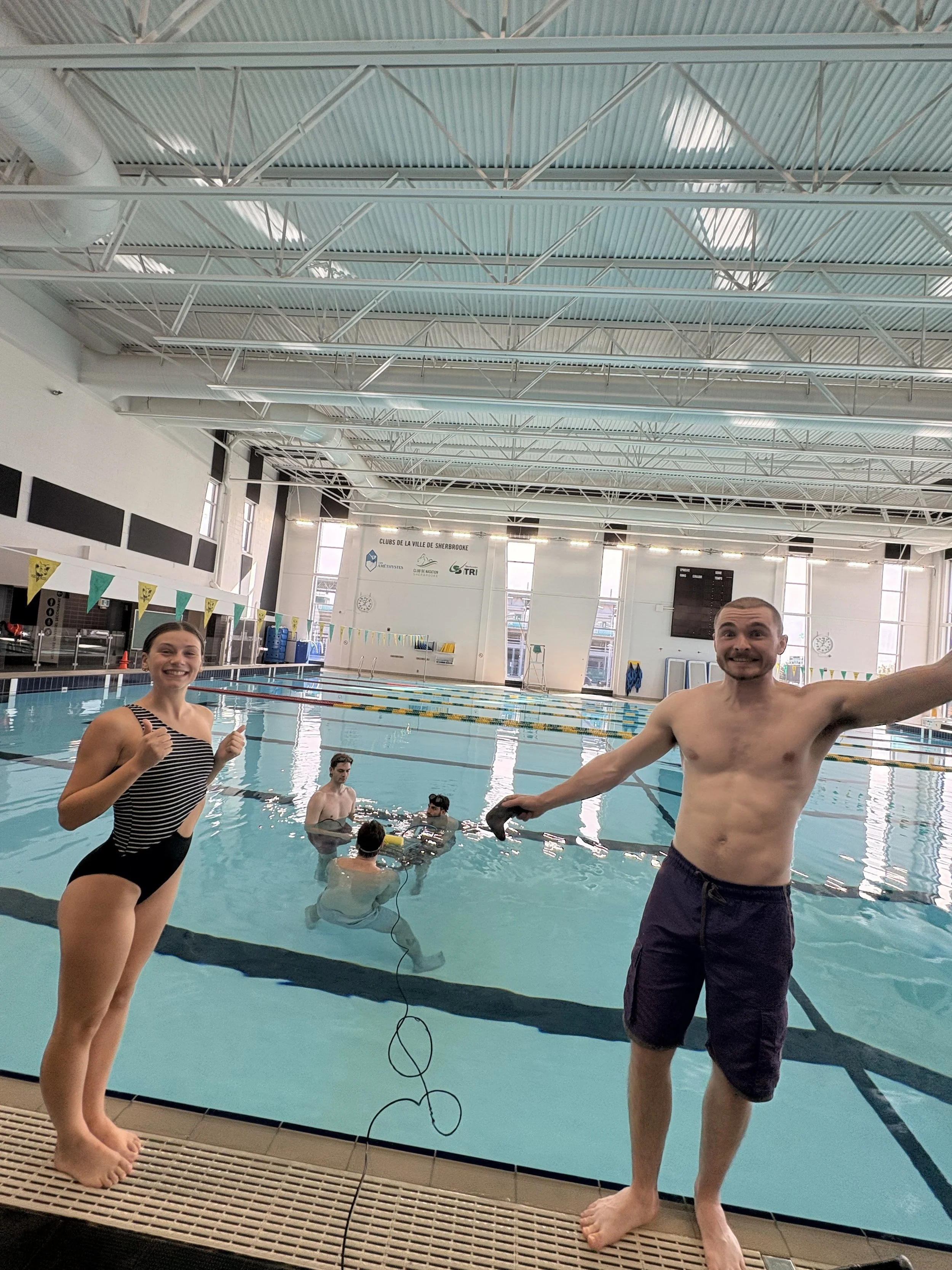 People at an indoor swimming pool, some in the water and others on the deck, with high ceilings and large windows in the background.