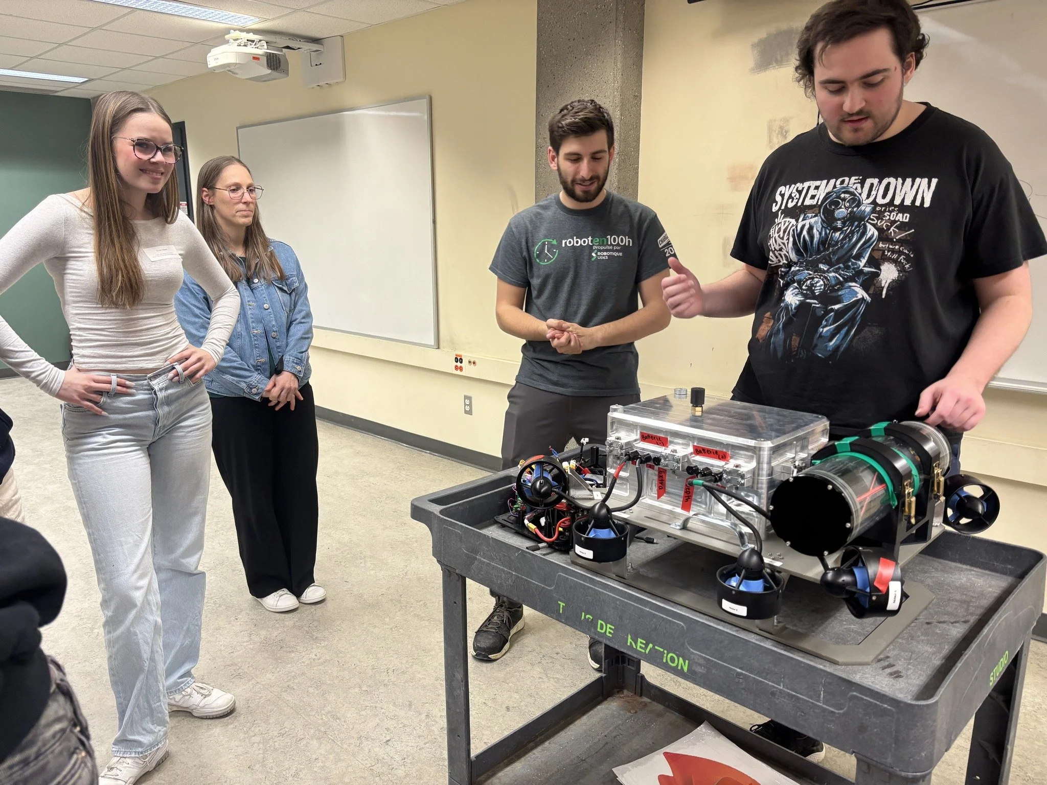 Four people observe a robotics project on a wheeled table in a classroom. The project includes a clear plastic box with labeled batteries and various electronic components. One person points at the project while others watch.
