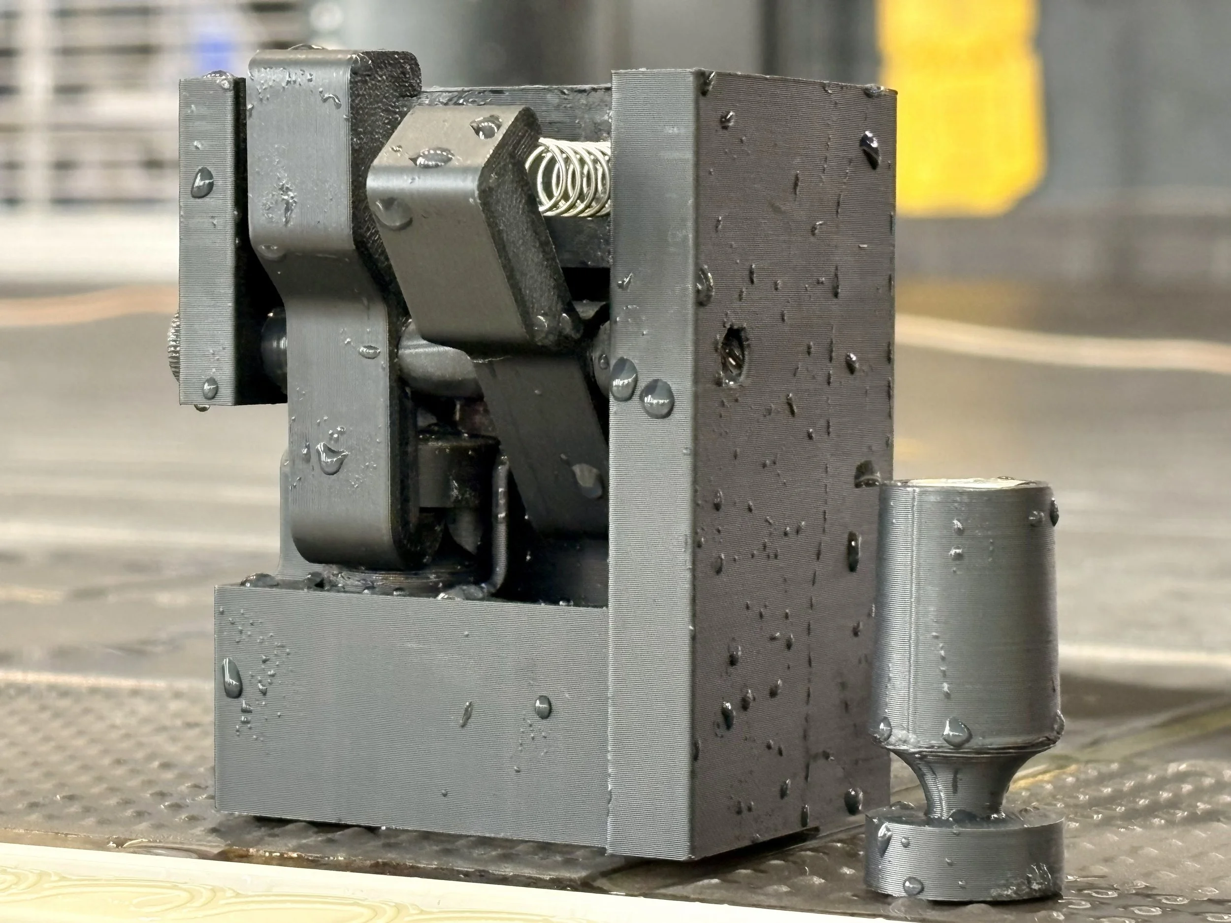 A close-up of a metal industrial machine component with water droplets on its surface, situated on a flat surface, with a blurred background including a yellow object.