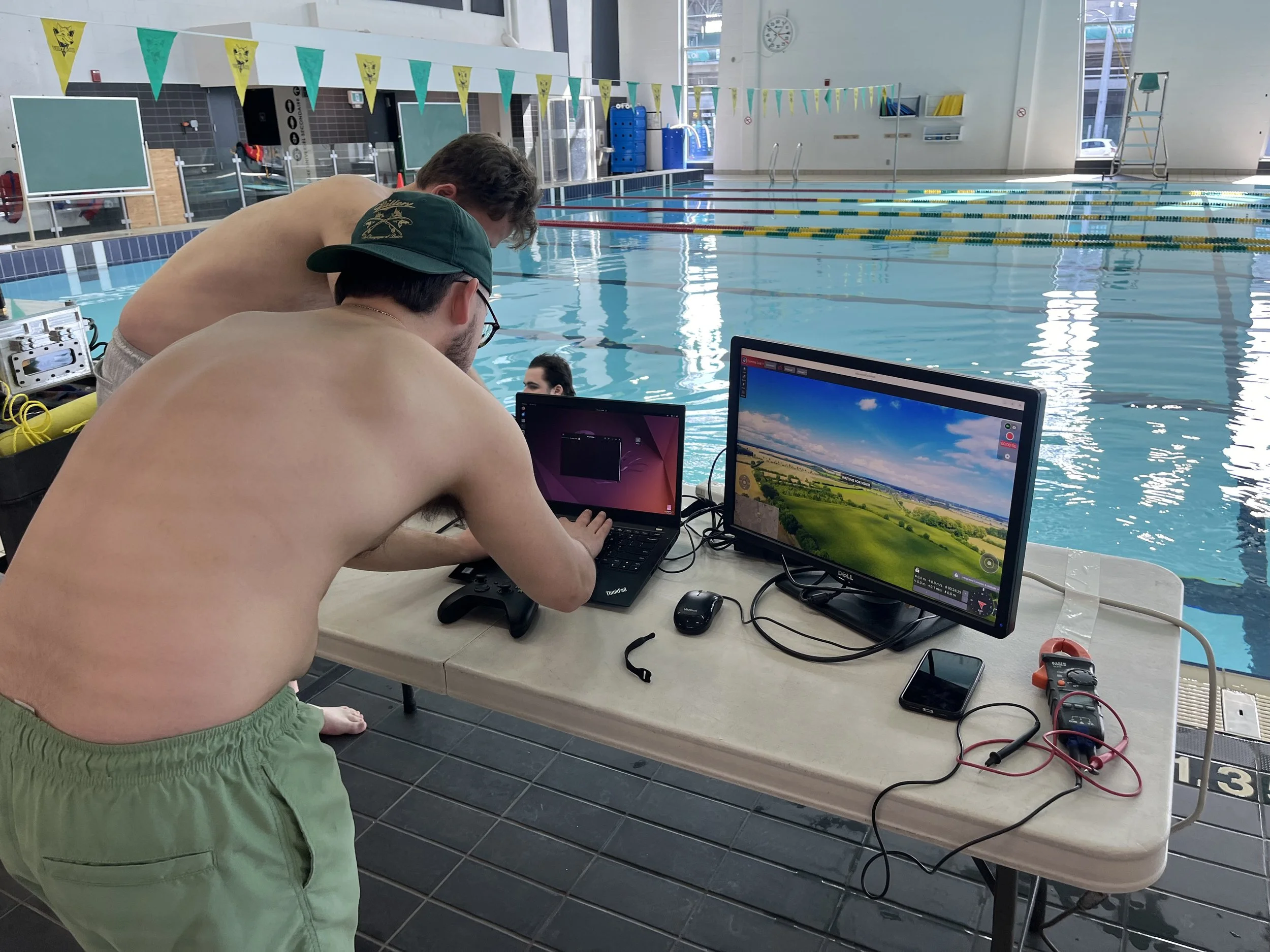 An indoor swimming pool with two prisoners sitting next to each other in swimsuits, appearing to be working on technology and filming. There are two laptops and various electronic equipment on a table near the pool.