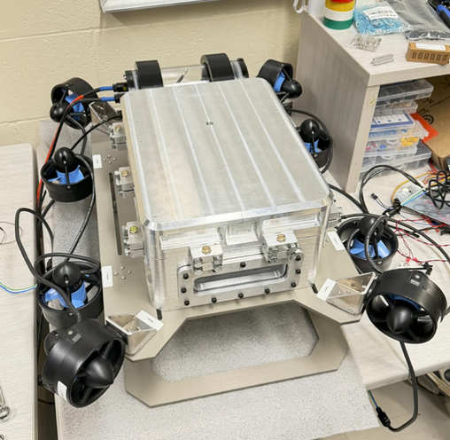 A robot with six black spherical wheels arranged around a central metallic rectangular body, on a table in a workshop or laboratory setting.