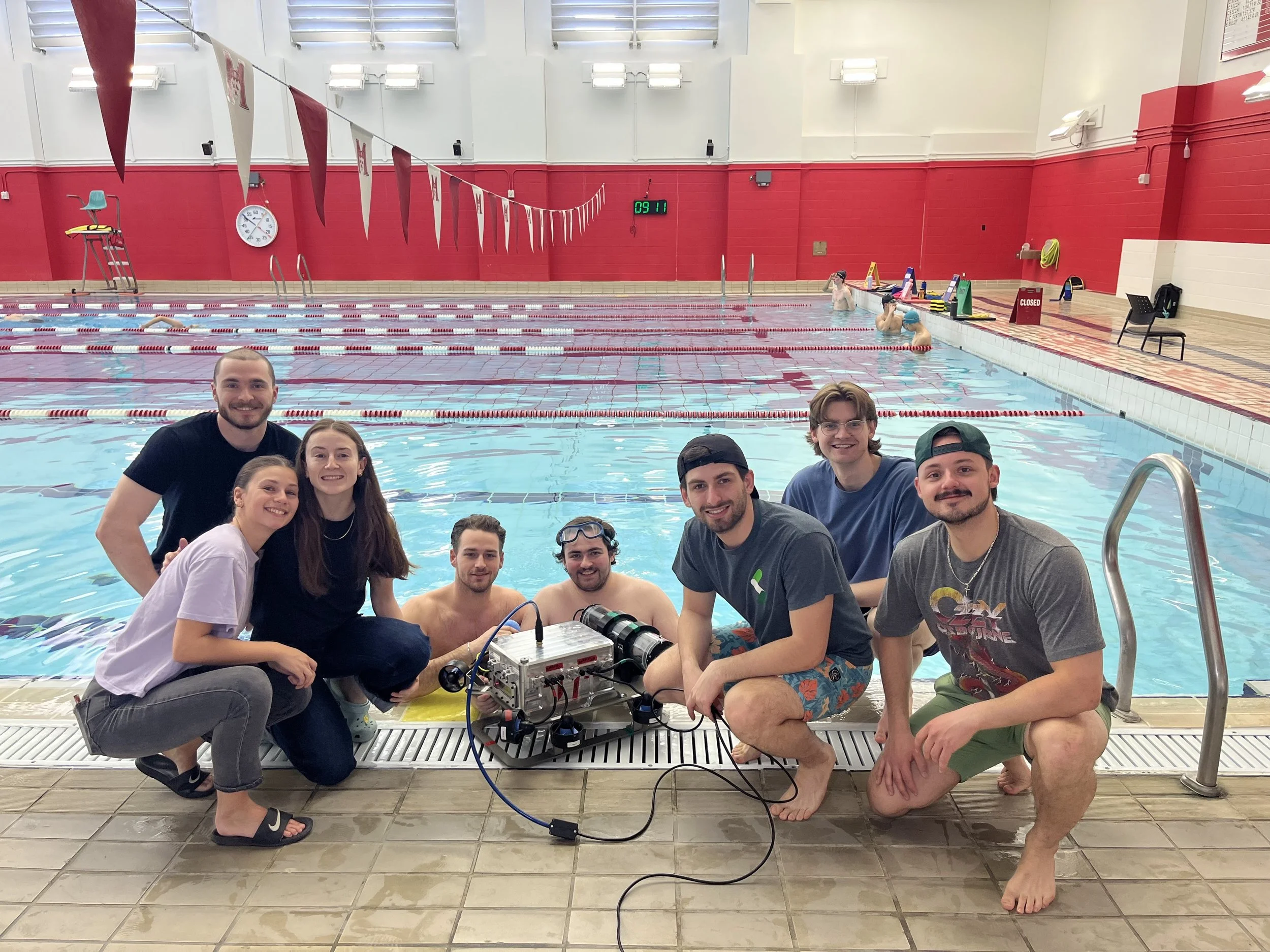 Group of nine people posing in front of an indoor swimming pool, with some individuals crouching and others standing, near a robotics project. The pool has red and white lane dividers, and there are swimmers in the water, some with goggles. The background shows a red and white wall, a large clock, and various swimming equipment and safety signs.