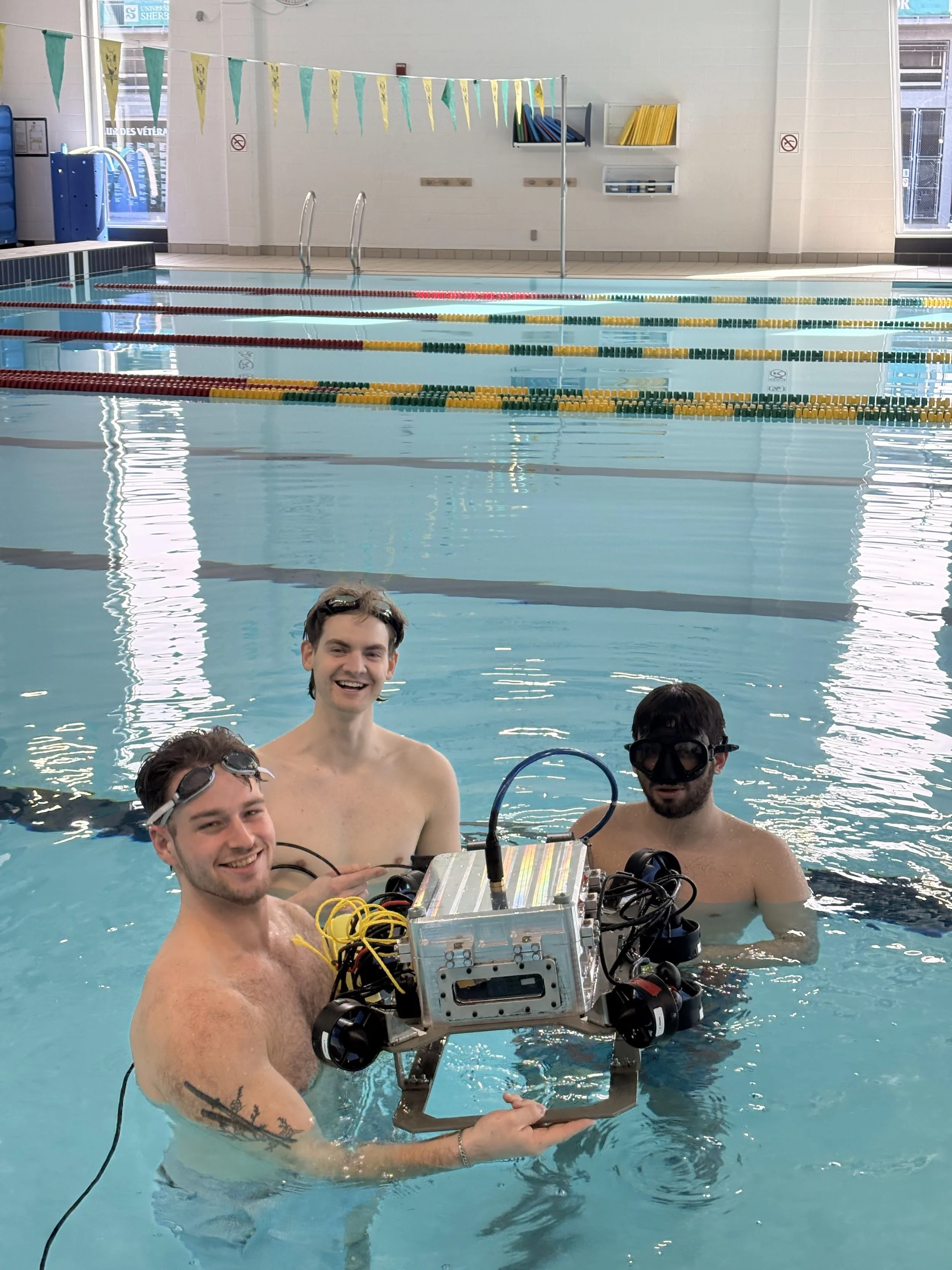 A group of four young men in a swimming pool holding an underwater robot with electronic components and wires. They are indoors, and one man is smiling at the camera while the others wear goggles. The background shows climbing stripes, pool equipment, and windows.