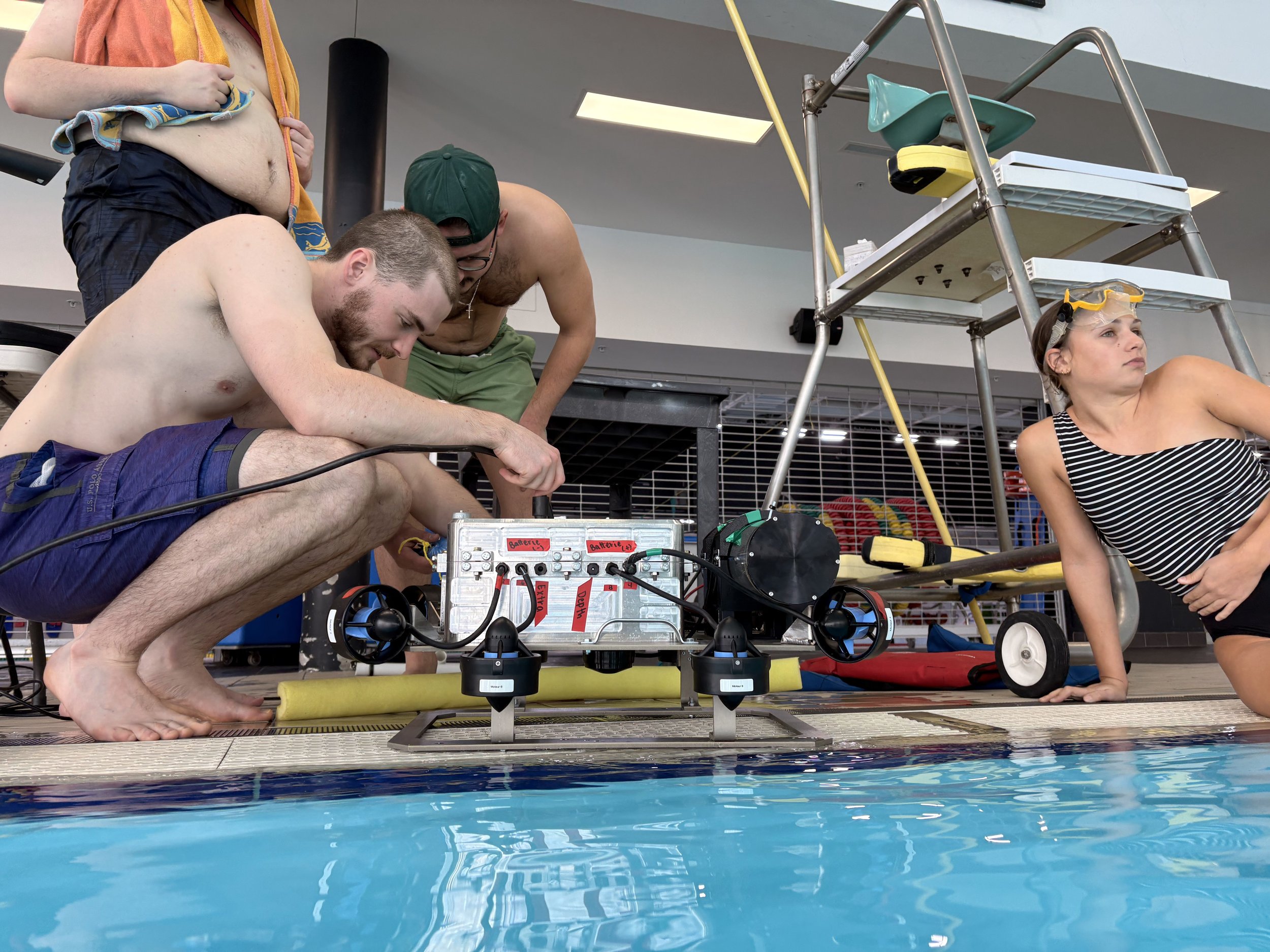 A group of people working on a robotic underwater drone next to a swimming pool, with one person wearing safety goggles and others observing.