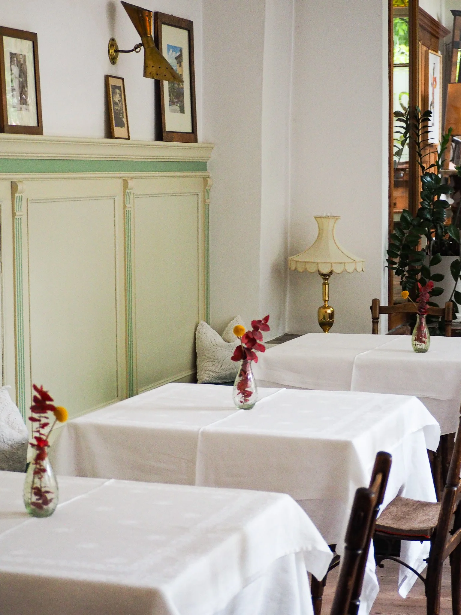 Gemütlich gedeckter Tisch mit weißen Tischtüchern, Gläsern mit Blumen, im Hintergrund ein altertümliches Möbelstück, Wandlampen und Bilder an der Wand, Fenster mit Blick nach draußen.