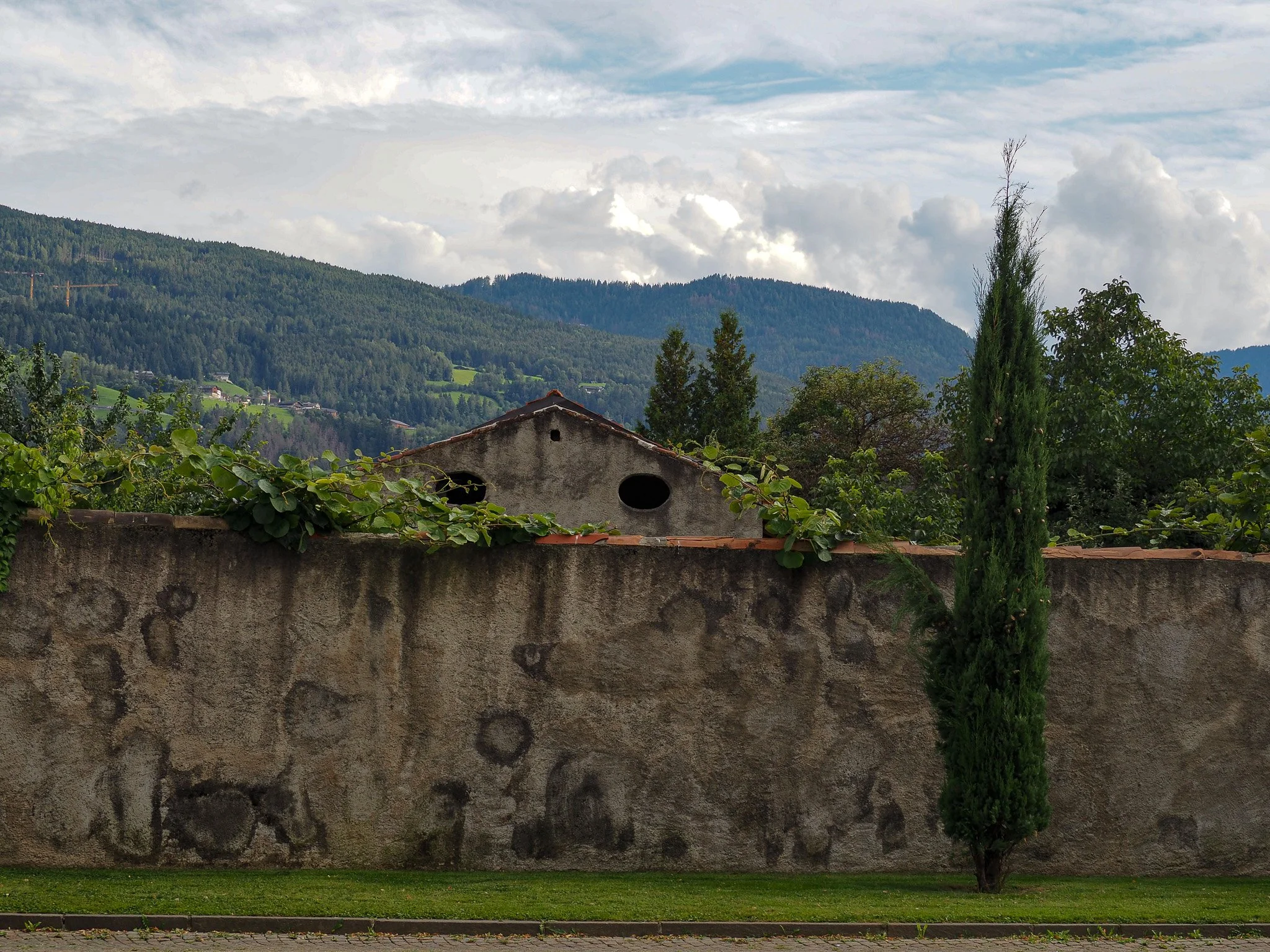 Eine alte Steinmauer mit grünem Bewuchs davor, im Hintergrund ein Gebäude mit runden Fenstern, und eine Landschaft mit Hügeln, Bäumen und Wolken am Himmel.