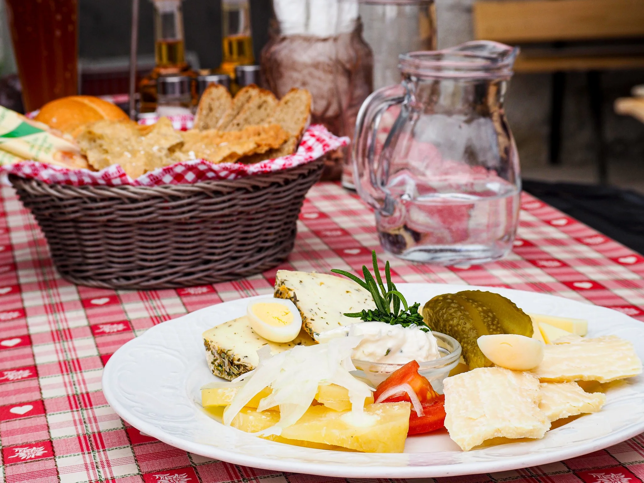 Ein Teller mit verschiedenen Käsesorten, eingelegter Gurke, Tomate, Zwiebel, Knoblauchzehe, Ei, frischen Kräutern und einem Glas Wasser auf einem rot-weiß karierte Tischdecke. Im Hintergrund ein Korb mit Brot und eine Wasserkrug.