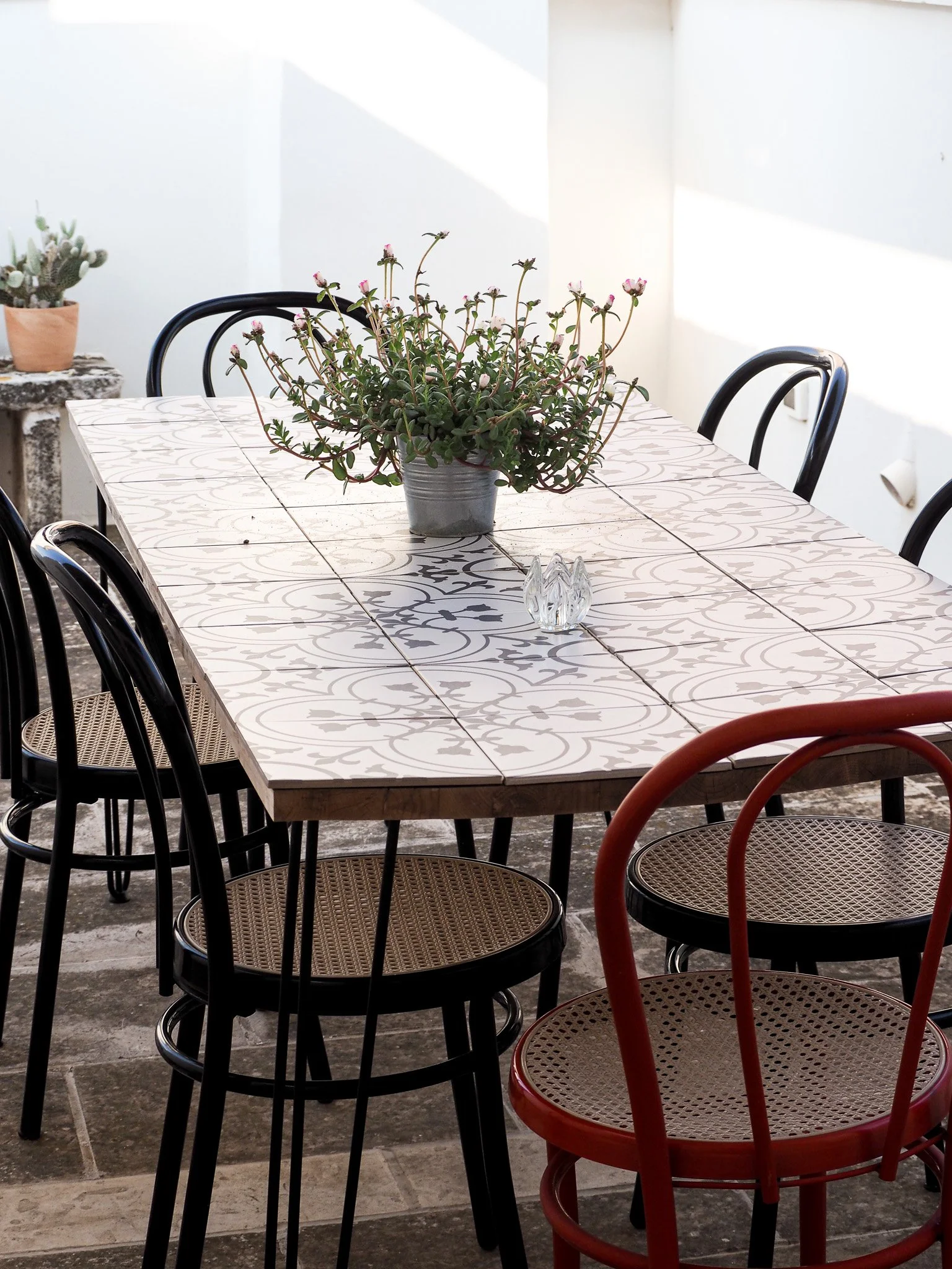 Ein rechteckiger Tisch mit weißen, bemalten Fliesen und schwarzem Rand, auf dem sich eine Topfpflanze und eine kleine Kristallvase befinden. Um den Tisch herum stehen Stühle in Schwarz, Rot und mit Rattanstühlen.