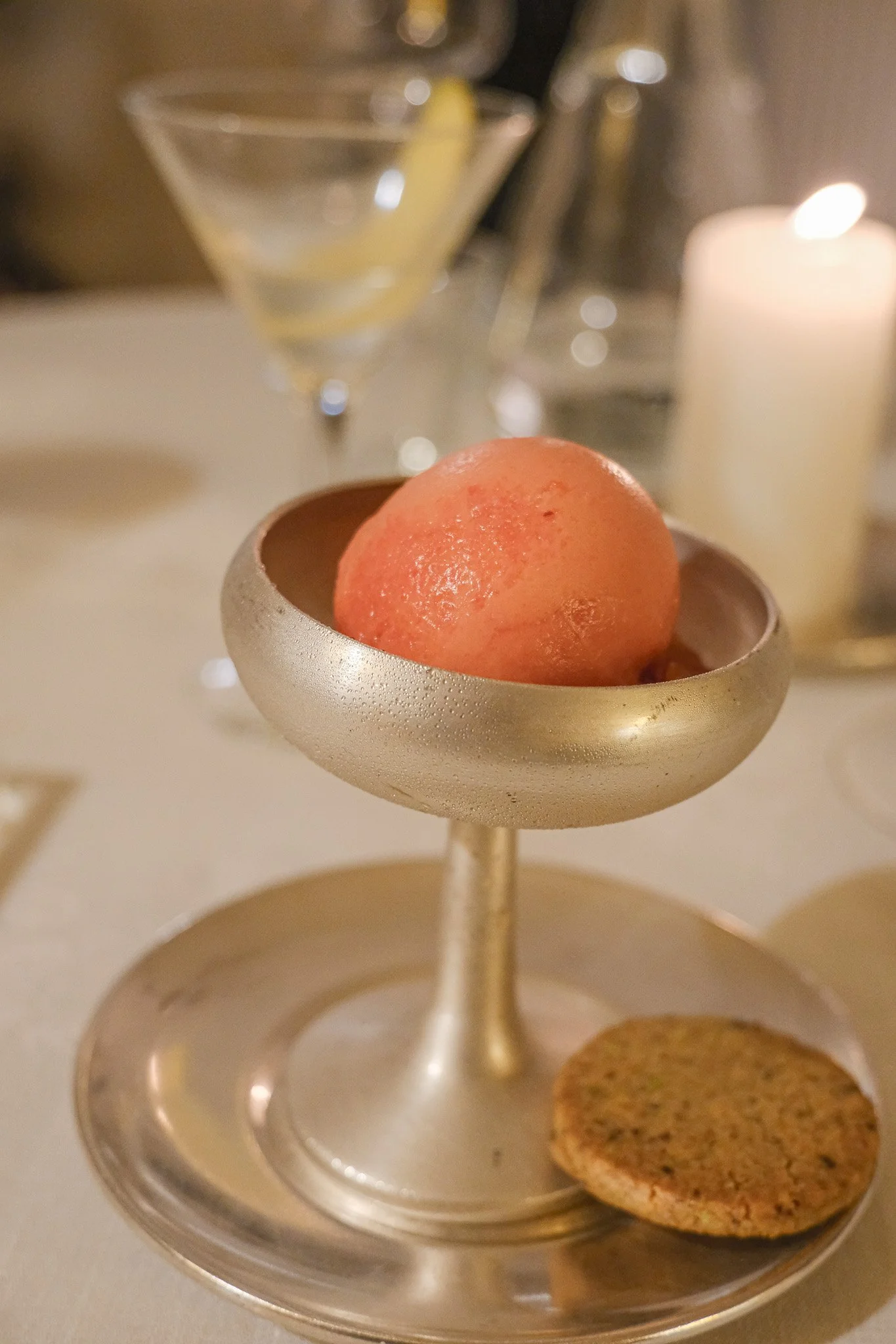 Ein goldener Stand mit einer roten Maracuja und einem Keks, im Hintergrund zwei Cocktailgläser und eine brennende Kerze.