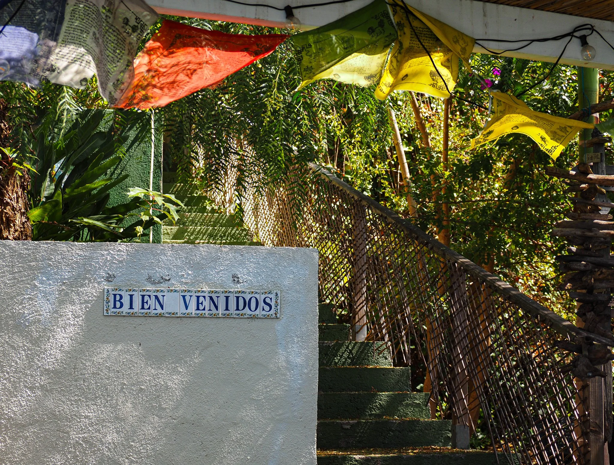 Ein weißer Wand mit einem blauen und weißen Fliesen-Schild, das 'BIENVENIDOS' geschrieben ist, und grüne Treppe, umgeben von Bäumen und Pflanzen, mit bunten Gebetsfahnen im oberen Bereich.