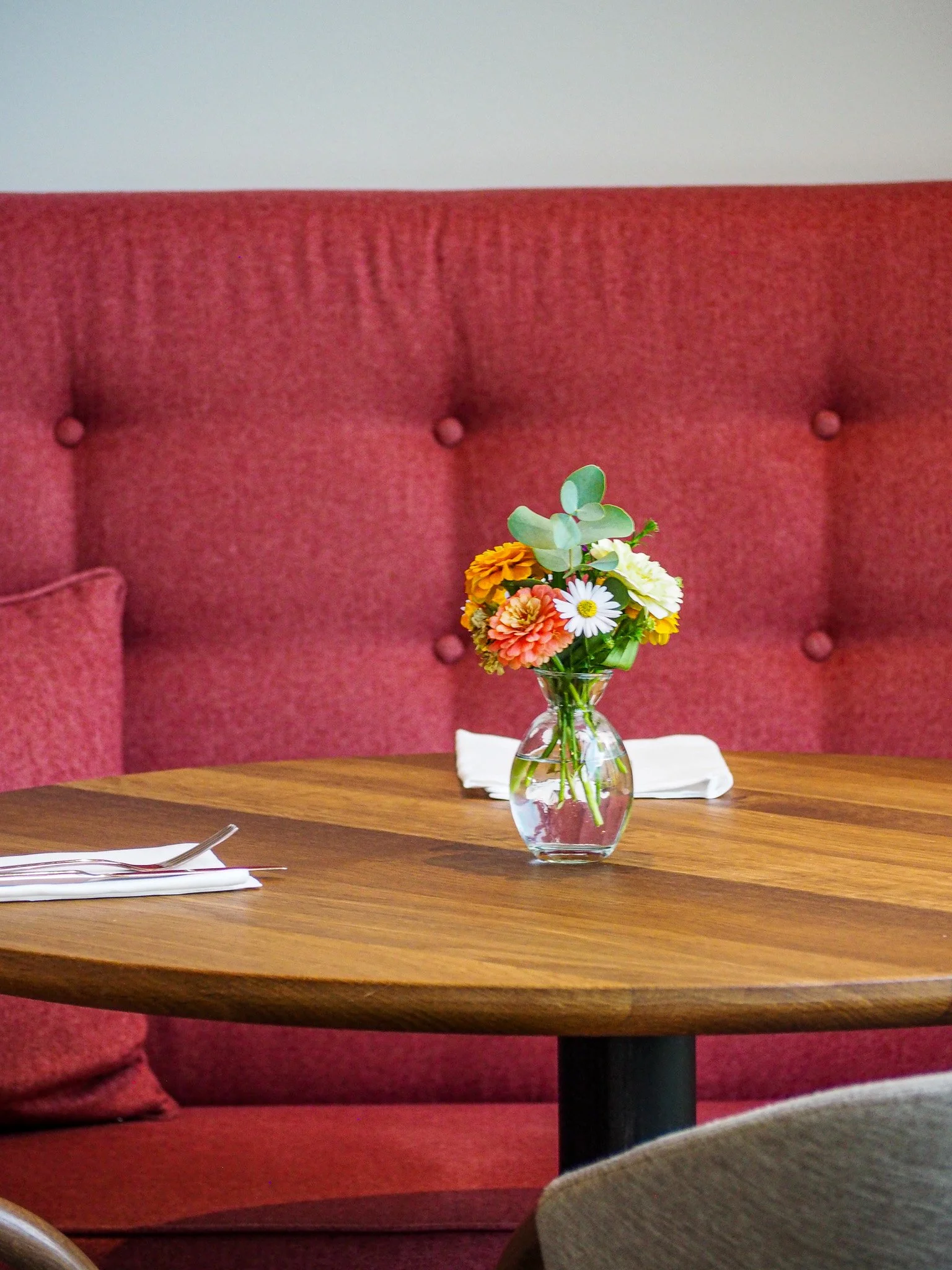 Ein Blumenarrangement mit bunten Blumen in einer klaren Vase auf einem Holztisch in einem Raum mit roter Polstermöbel.
