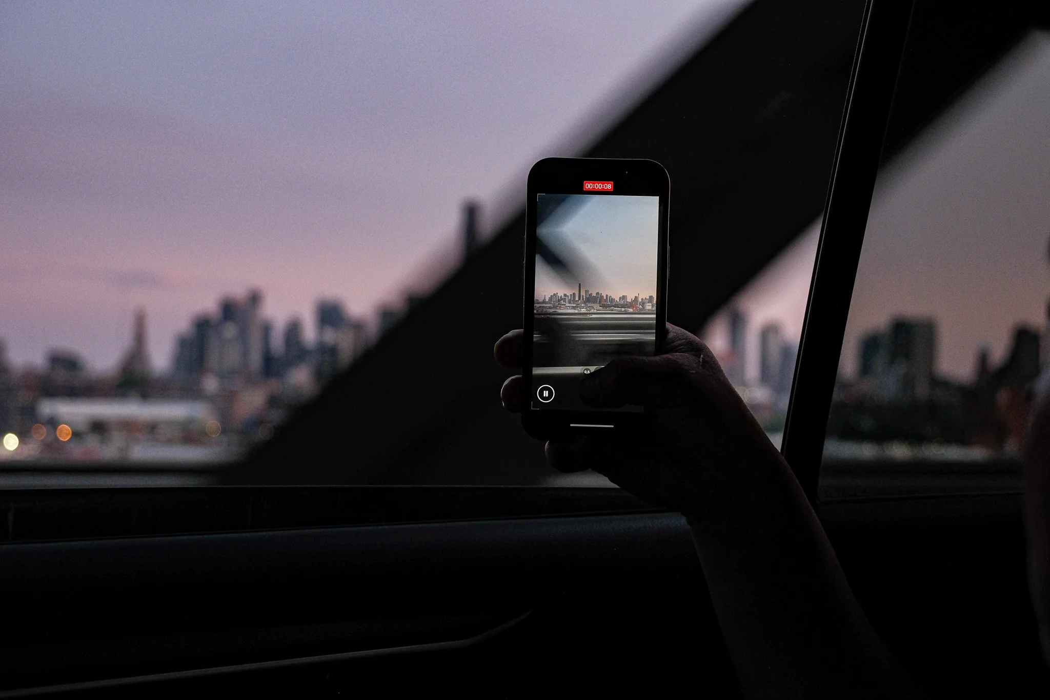 Eine Person filmt die Skyline einer Stadt bei Sonnenuntergang mit einem Smartphone durch ein Autofenster.