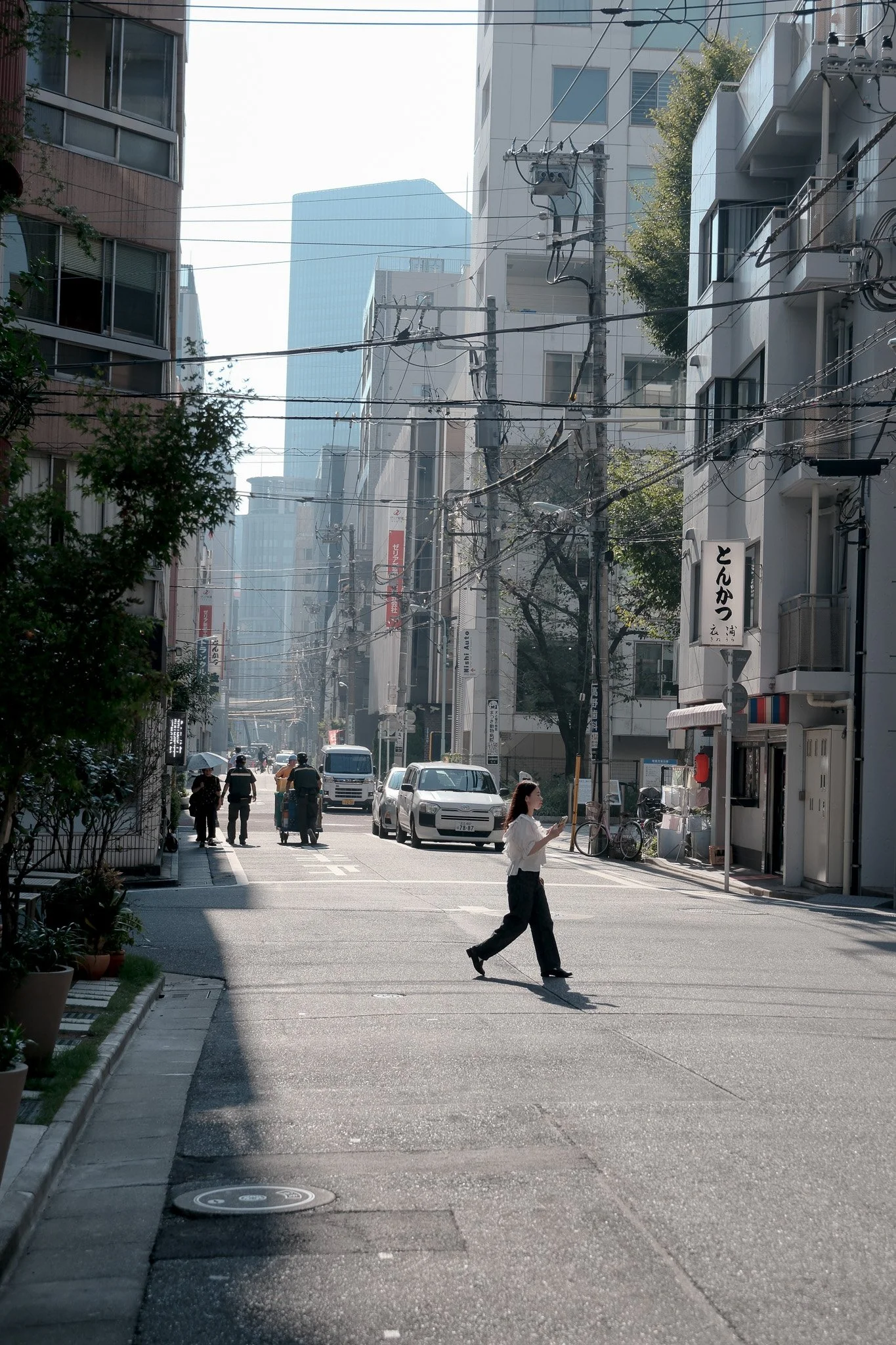 Stadtstraße in Japan mit Hochhäusern, mehreren Fußgängern, Autos und Stromleitungen, eine Frau geht über die Straße, es ist sonnig.