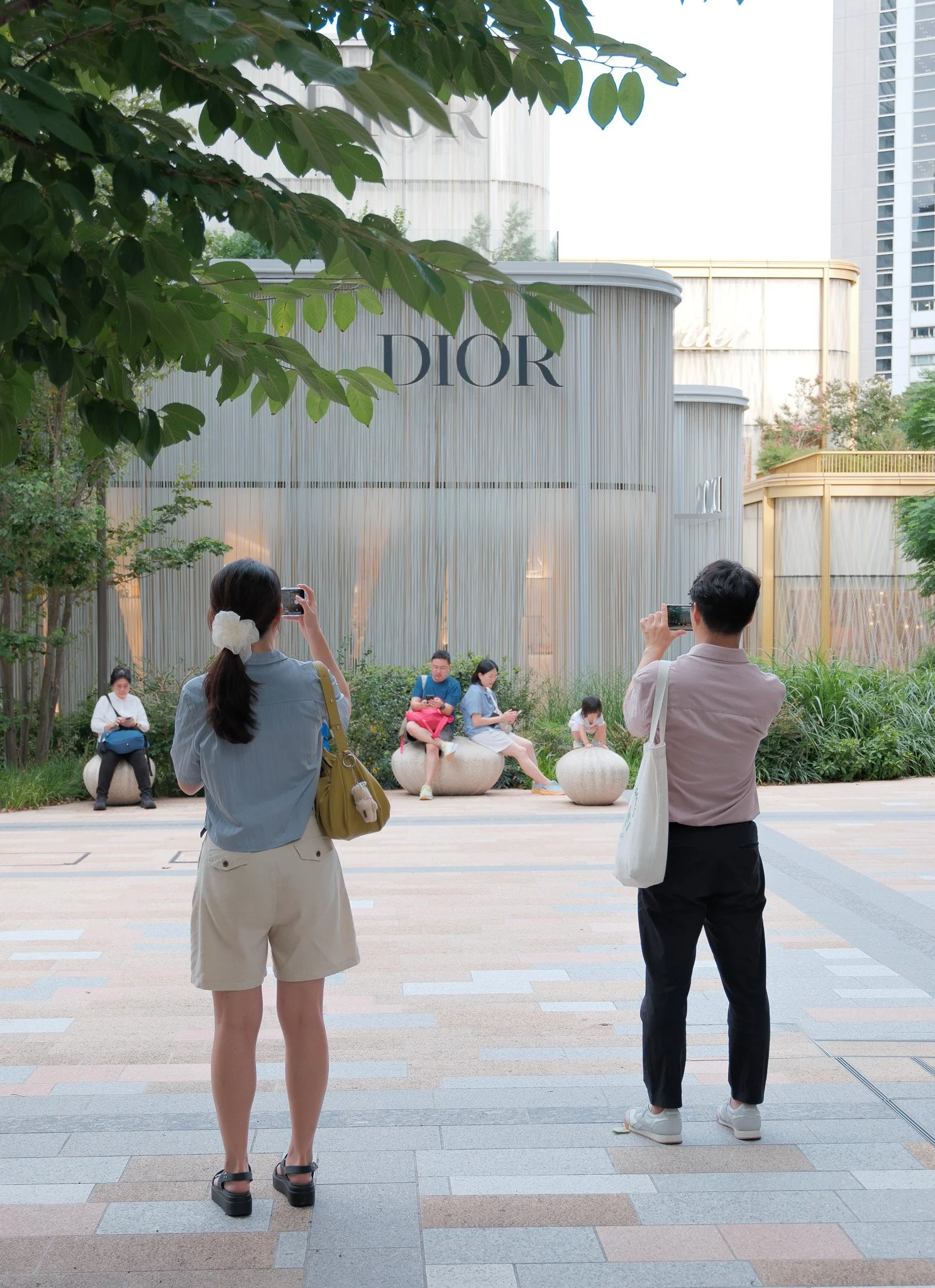 Zwei Menschen fotografieren eine Gruppe von Personen, die auf Sitzflächen vor der Dior-Filiale sitzen, mit großen Gebäuden im Hintergrund.