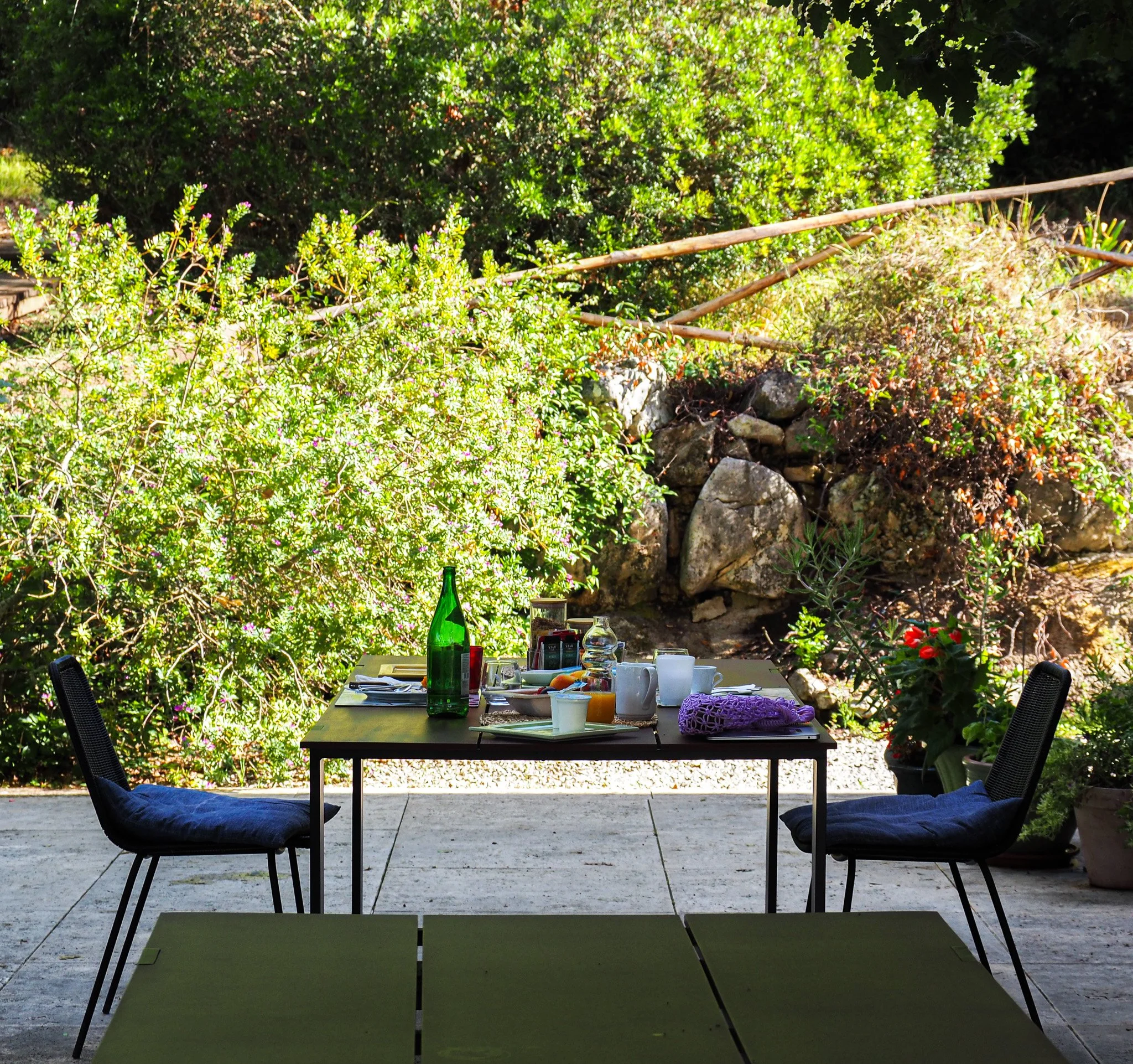 Ein Frühstückstisch im Freien mit Tassen, Flaschen, Besteck und Dekoration, vor einer grünen Gartenzone mit Pflanzen, Steinen und Bäumen.