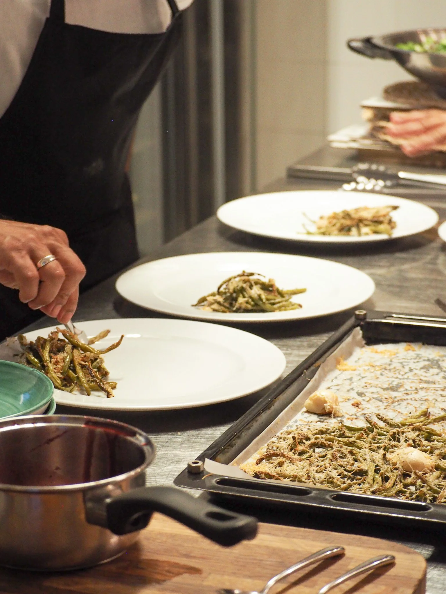 Koch bereitet mehrere Teller mit grünen Bohnen-Gratin vor, auf einem Küchenarbeitsbereich, mit einem Backblech und Kochutensilien im Vordergrund.