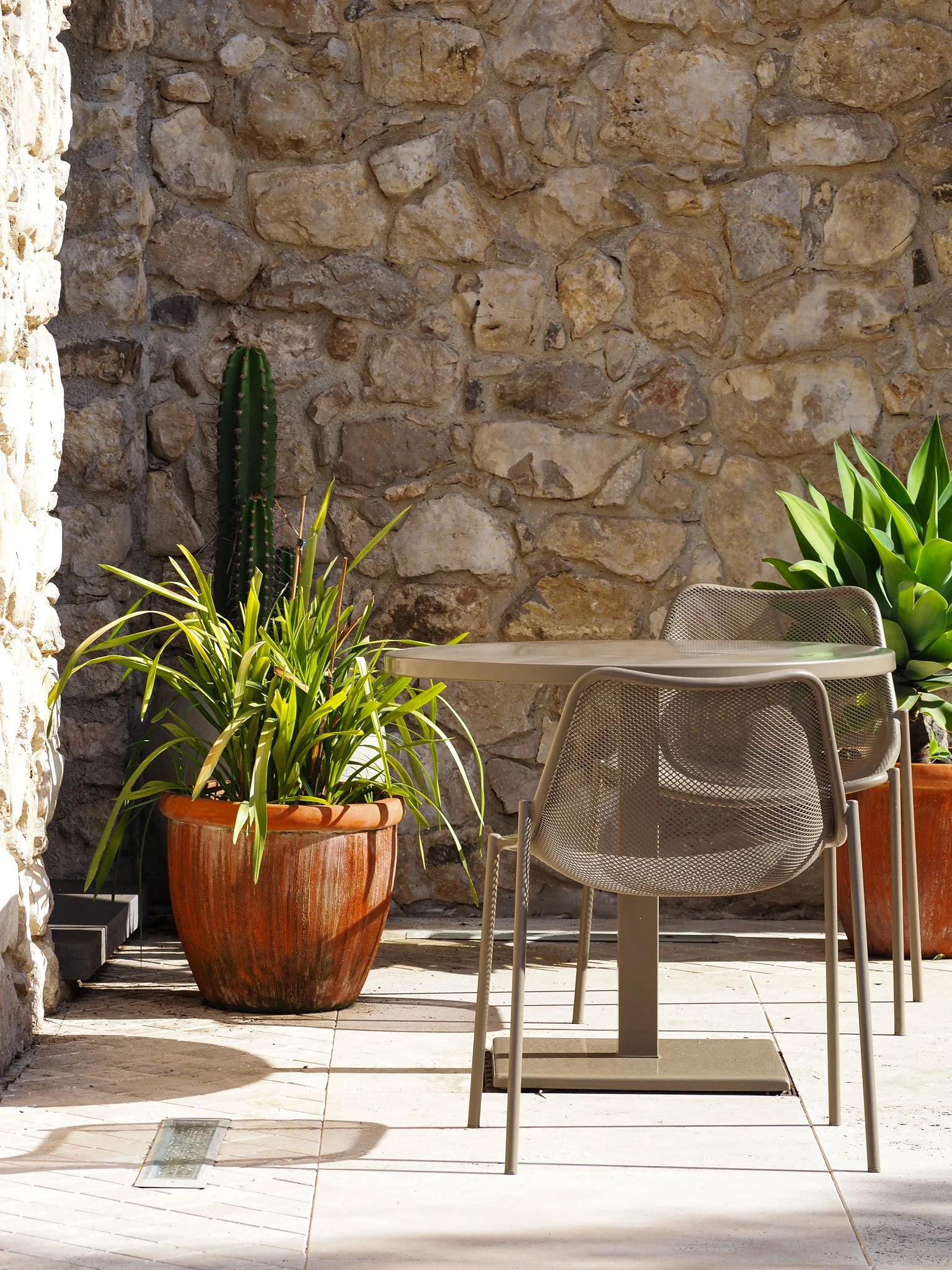 Ein Terrasse mit einem Steingarten, zwei große Tontöpfe mit Pflanzen, ein Tisch und sechs Klappstühle, an einer Steinmauer im Sonnenlicht.