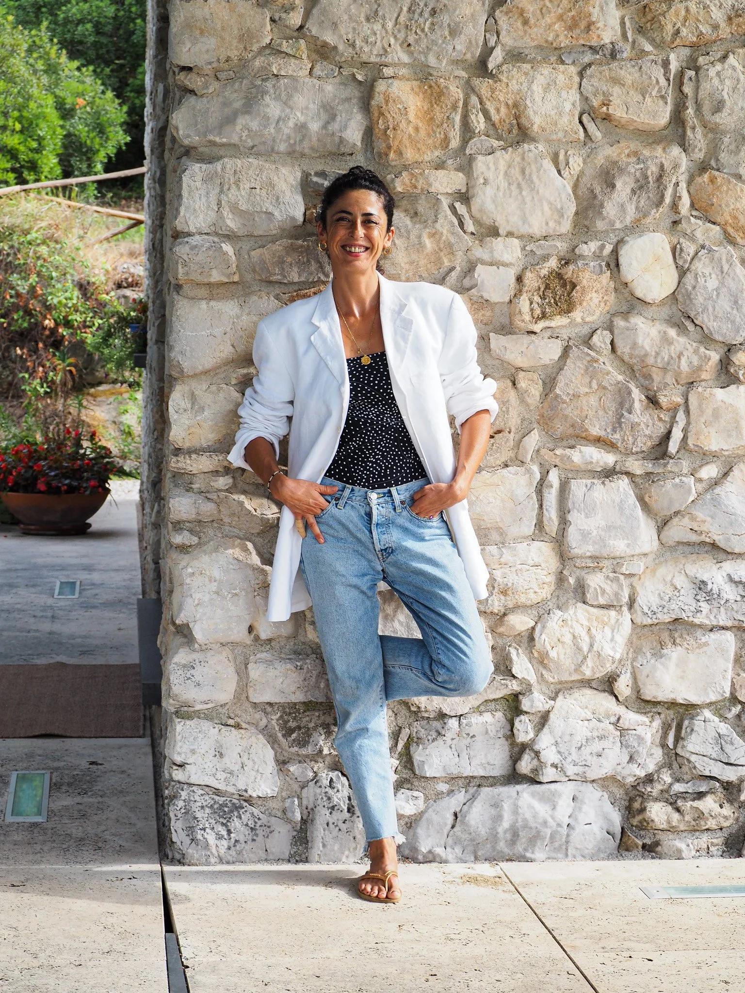 Frau steht am Steinmauer, trägt weiße Jacke, schwarzes Top mit weißen Punkten und Jeans, lacht.