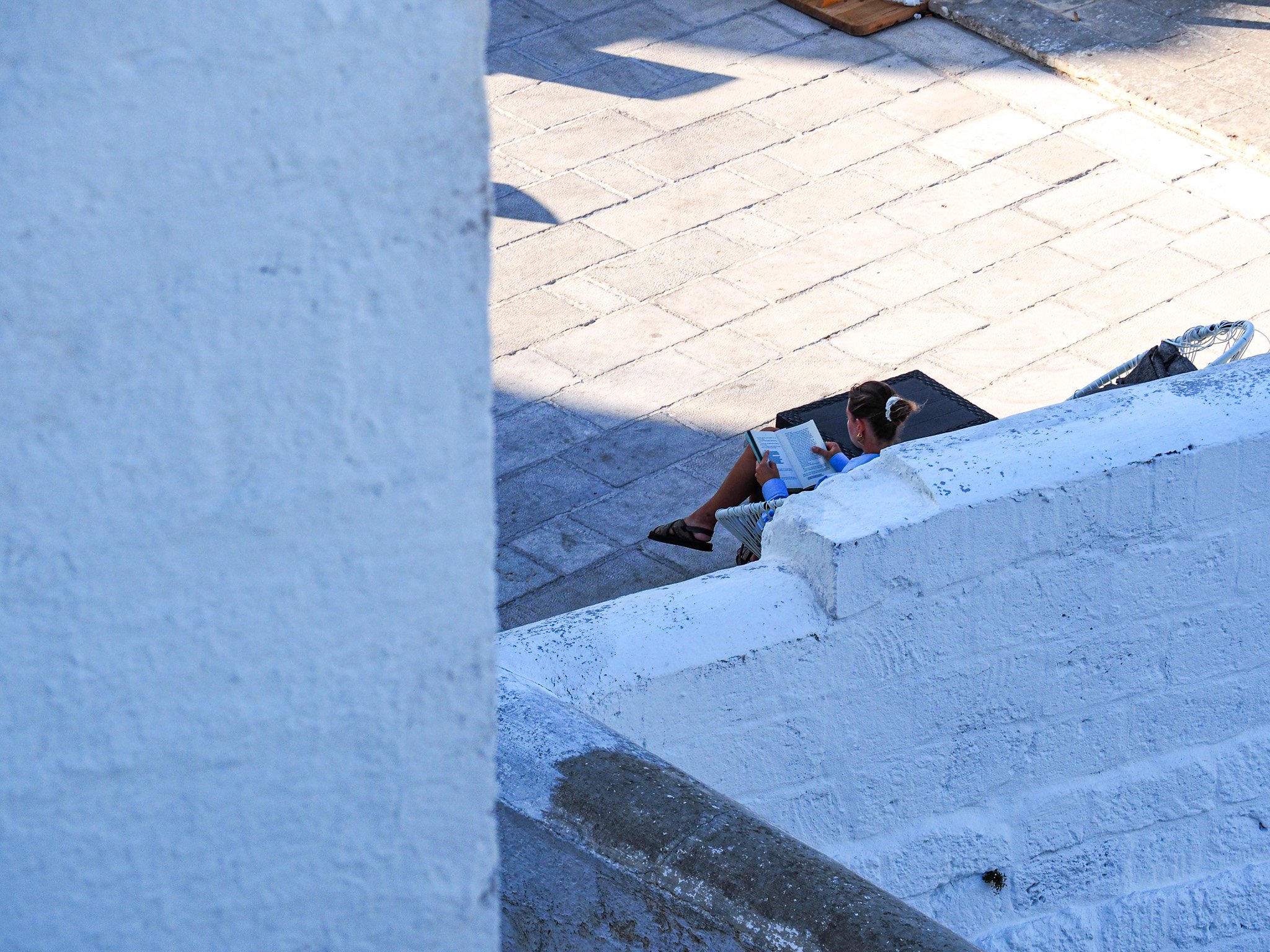 Eine Frau liest ein Buch, sitzt auf einer weißen Mauer im Freien, neben einer weißen Wand, auf gepflastertem Boden.