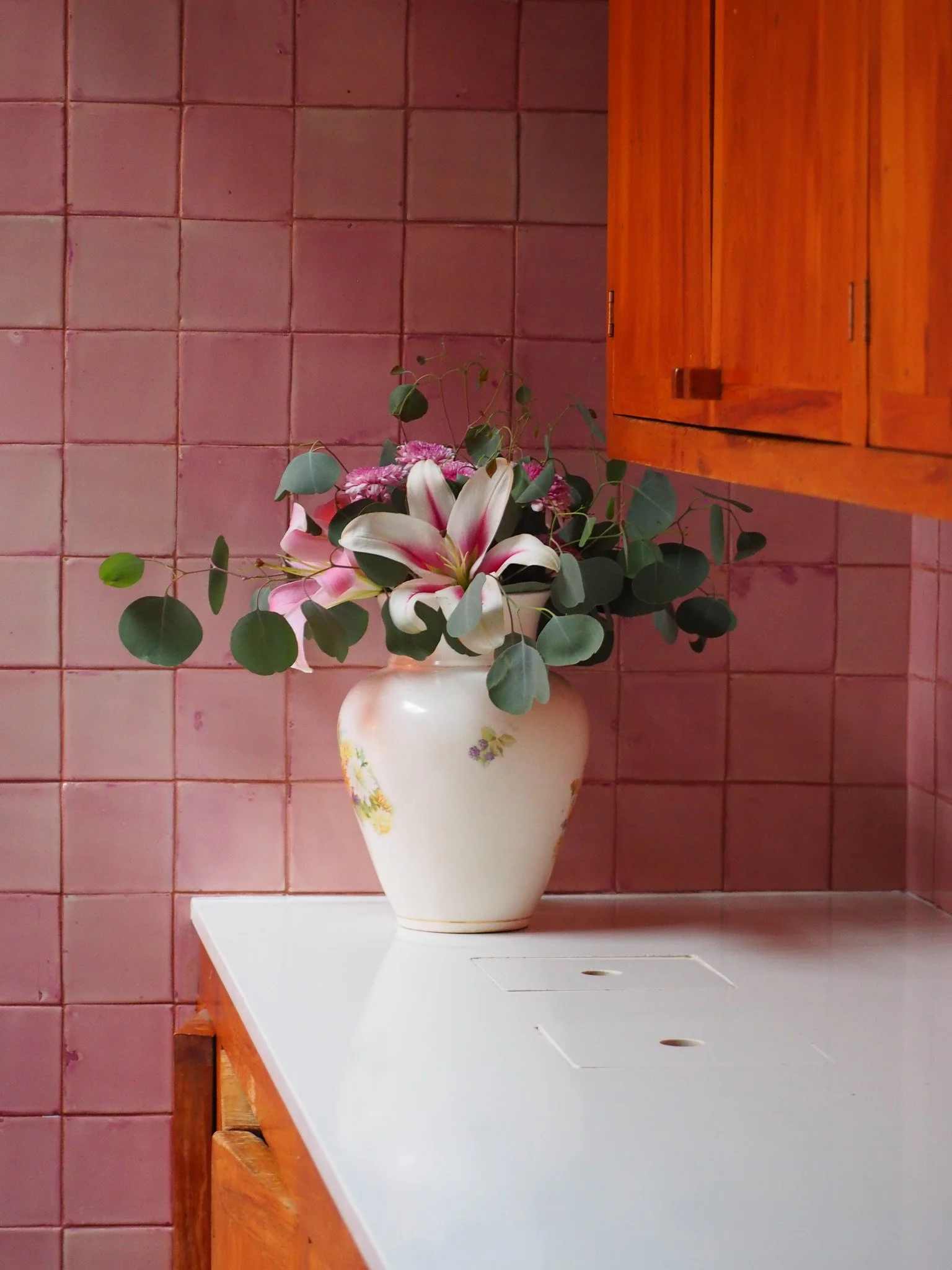 Blumenstrauß mit weißen und rosa Lilien in einer weißen Vase auf einer weißen Küchentheke vor rosa Fliesenspiegel.