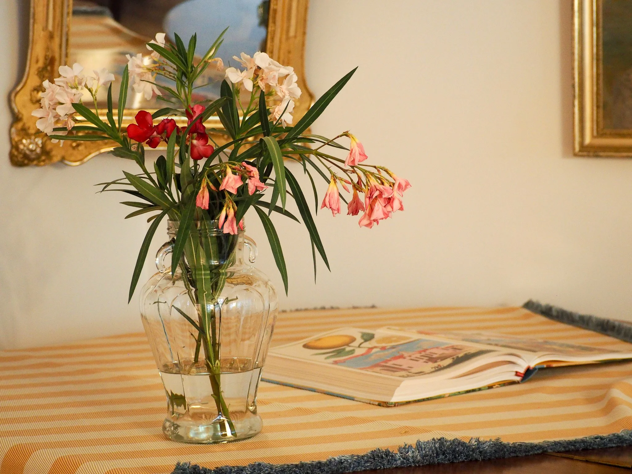 Blumenstrauß mit weißen, roten und rosa Blumen in einer Glasvase auf einem Tisch mit gelb-weiß gestreiftem Tuch, im Hintergrund eine goldgerahmte Spiegel und ein offen liegendes Buch.