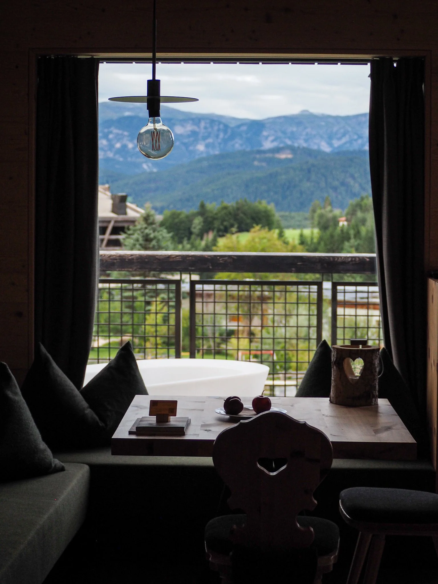 Blick aus einem Fenster mit Vorhängen auf eine Berglandschaft und einen Balkon mit Sitzgelegenheit, einem Bathtub im Hintergrund, einem Tisch mit Äpfeln und Dekorationen, eine Glühbirne hängt von der Decke.