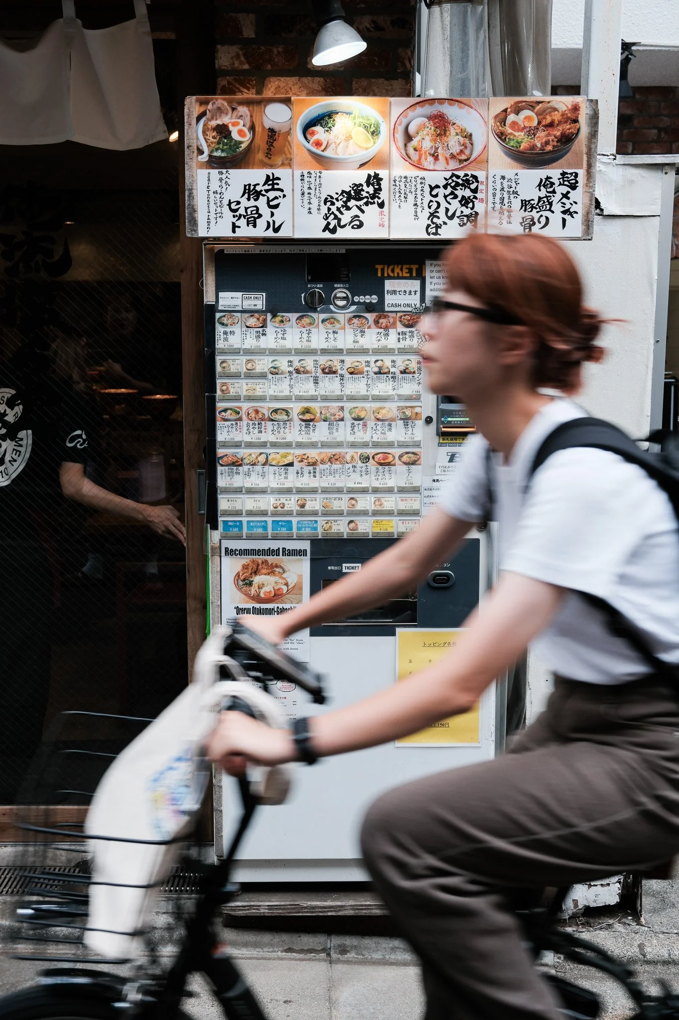 Frau auf Fahrrad fährt vor einem japanischen Restaurant mit Speisekarte und Bildern von Nudelsuppen