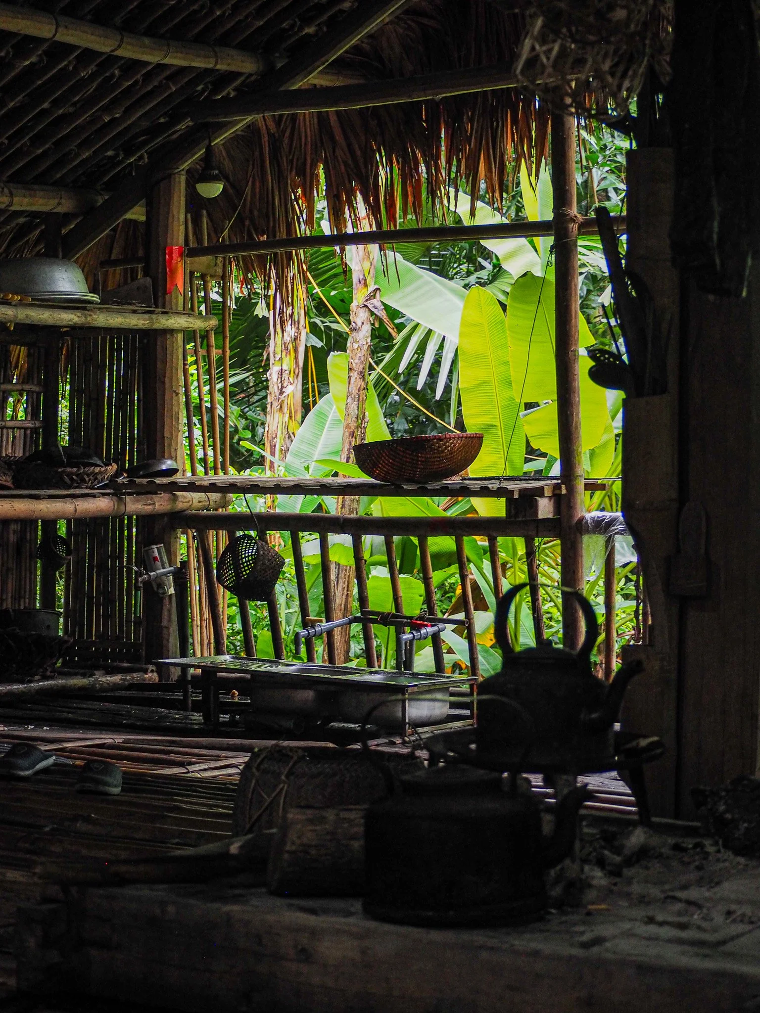 Innenaufnahme einer kleinen holz- und bambushütte mit Blick nach draußen auf grüne tropische Pflanzen, inklusive Bananenblätter, und einer offenen Küche mit Kochutensilien.