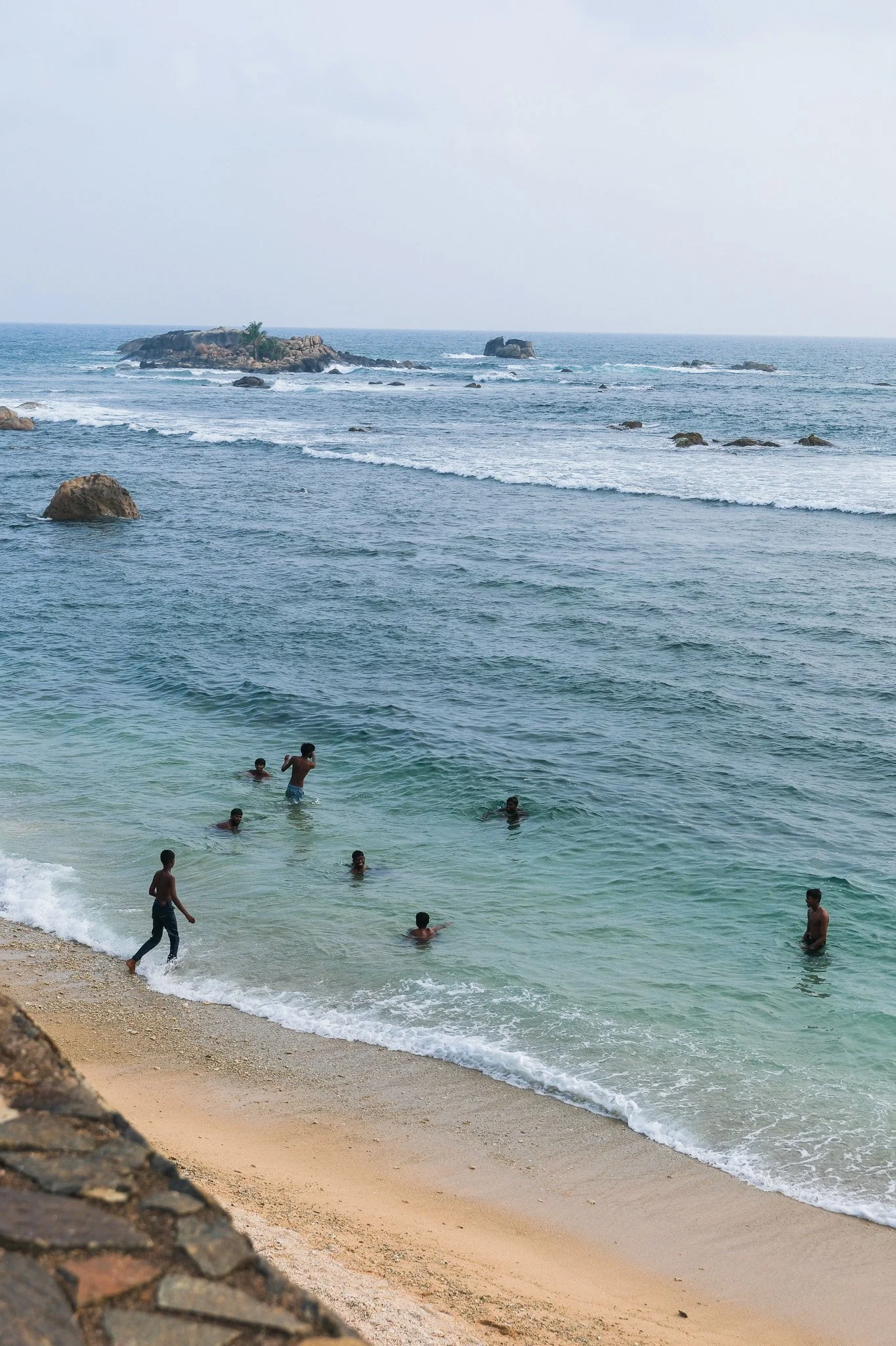 Mehrere Kinder und Jugendliche spielen und schwimmen im Meer an einem Sandstrand mit Felsen und kleinen Inseln im Hintergrund.