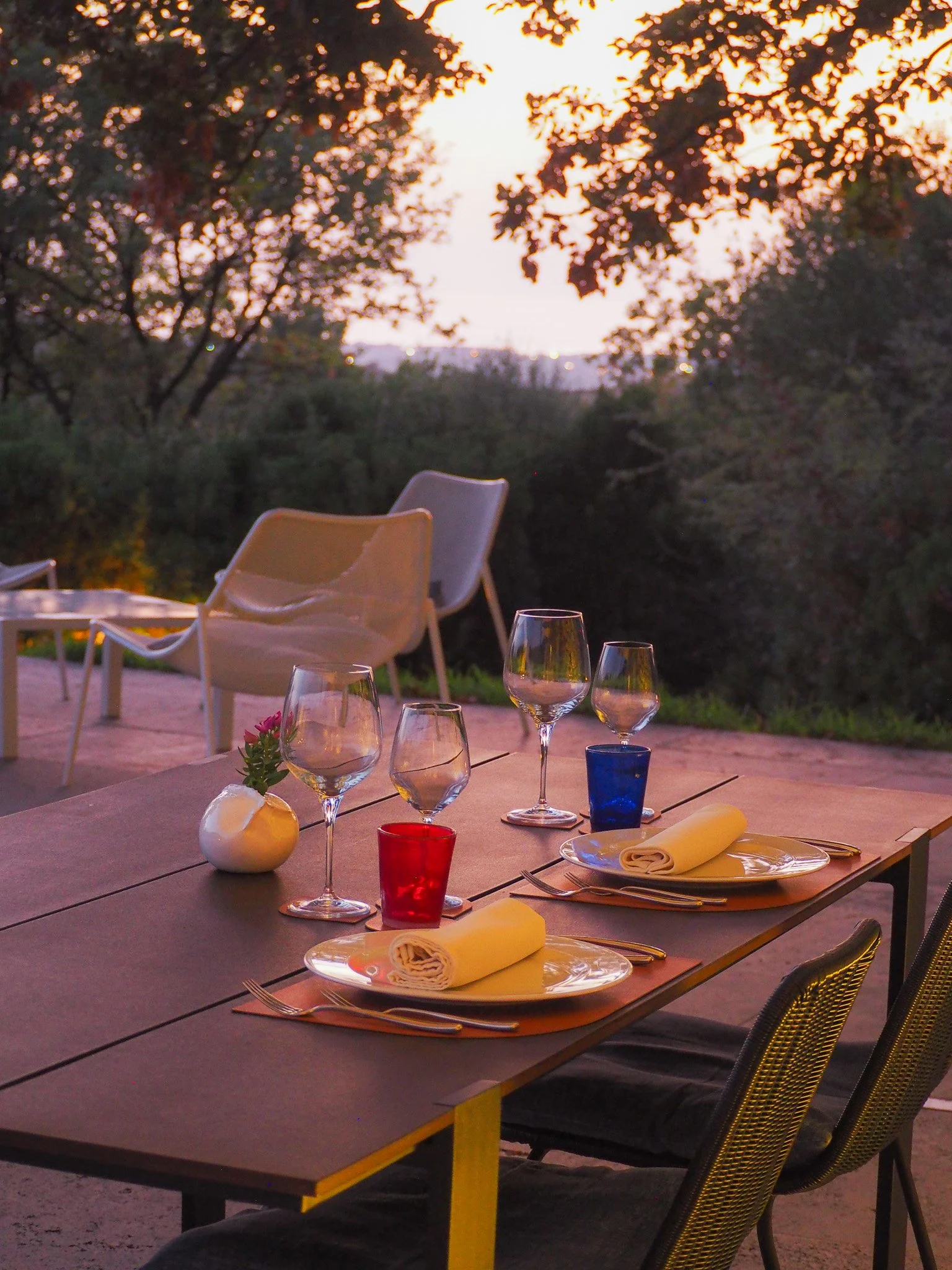 Ein im Freien gedeckter Esstisch mit mehreren Weingläsern, bunten Gläsern, weißen Tellern mit gedeckter Besteck, einer kleinen Vase mit Blume, bei Sonnenuntergang mit bewaldeter Umgebung im Hintergrund.