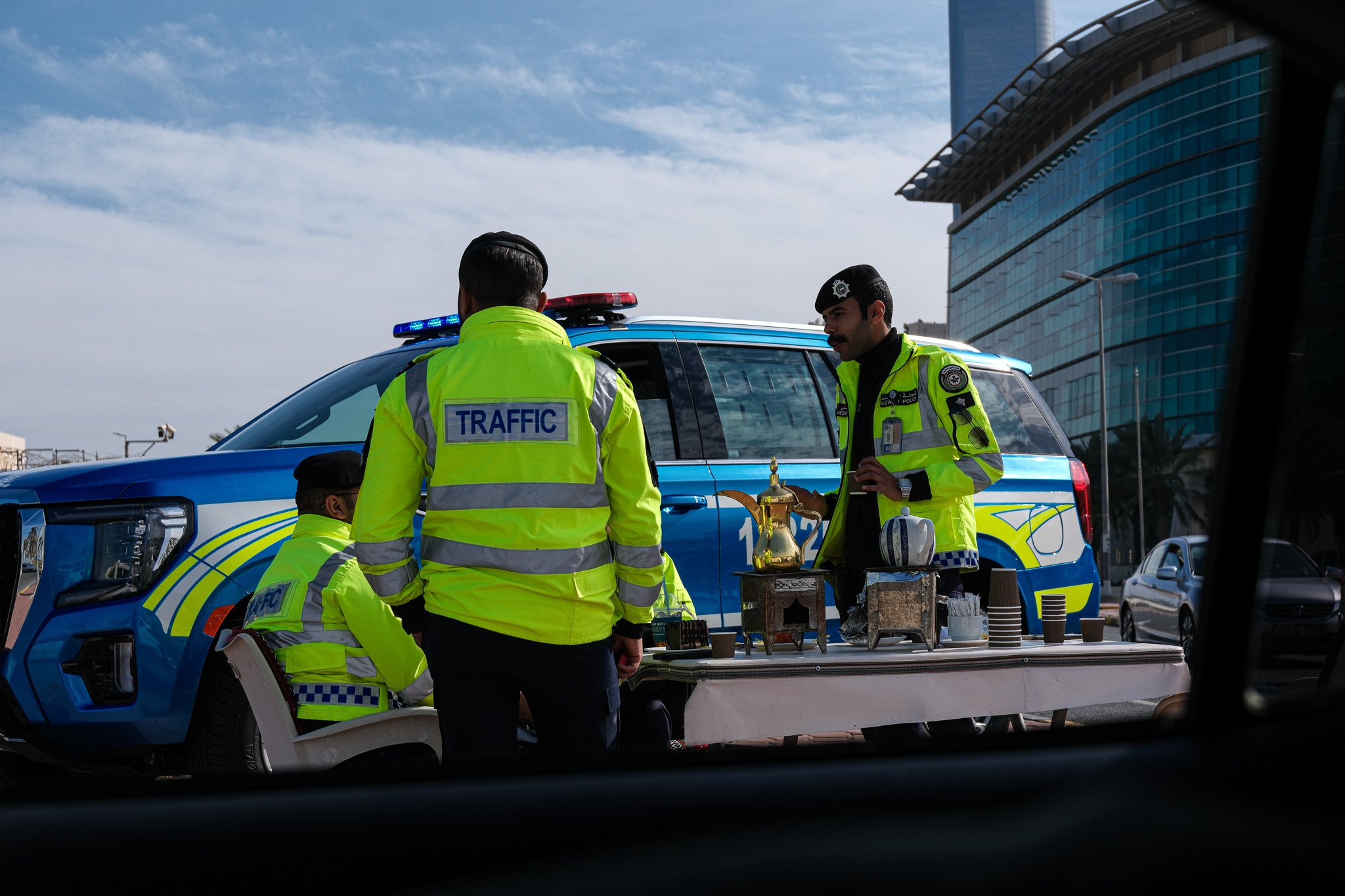 Drei Verkehrsbeamte in Neon-Gelben Jacken mit Aufdruck 'TRAFFIC' stehen an einem Tisch mit Kaffee und einer Kanne, vor einem blauen Polizei-Wagen mit blauem Licht auf dem Dach, im Hintergrund moderne Firmengebäude.