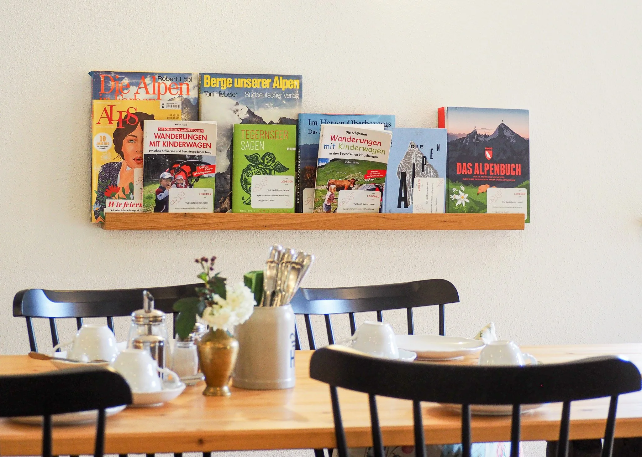 Ein Essbereich mit einem Holztisch, weißen Tassen, Tellern und Besteck. Auf dem Tisch steht eine Vase mit Blumen. An der Wand befindet sich ein Regal mit verschiedenen Fußbüchern und Reiseführern über die Alpen und Berge.