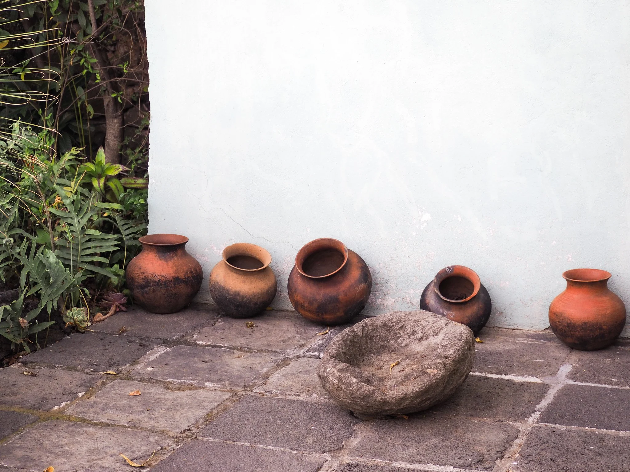 Fünf Terrakotta-Töpfe und ein großer Stein auf einem gepflasterten Boden vor einer weißen Wand.