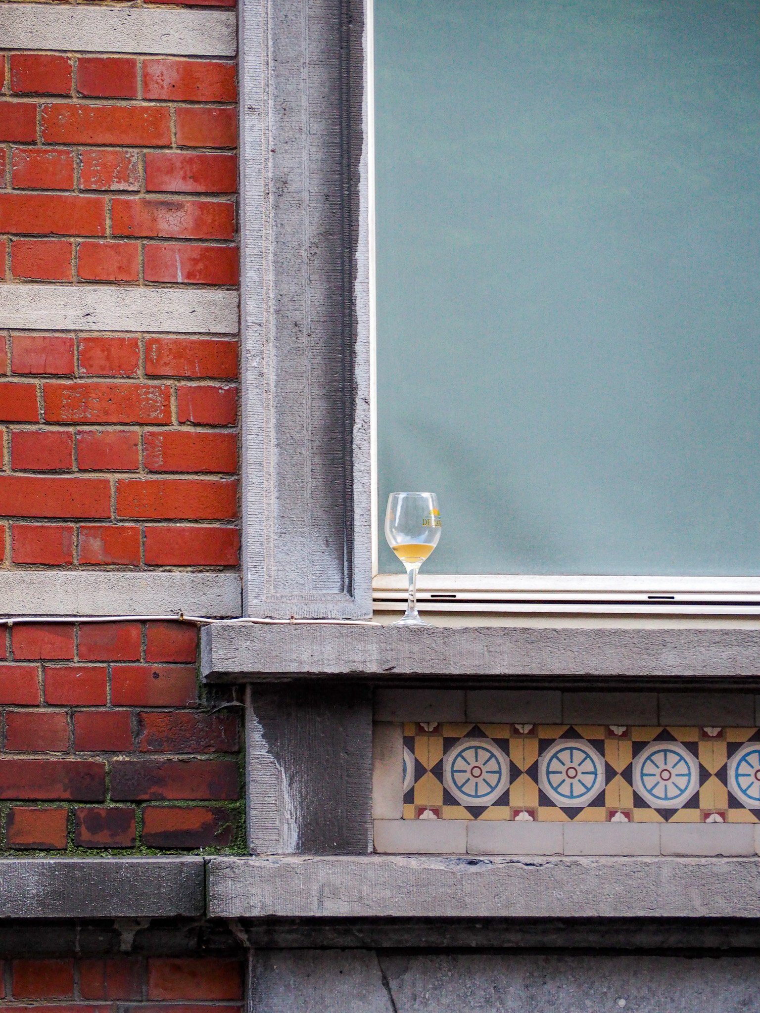 Ein leeres Weinglas mit einem kleinen Rest Wein steht auf einem Fenstersims vor einer grünen Fensterscheibe, neben einer Wand aus roten Ziegelsteinen und einem Holzrahmen.