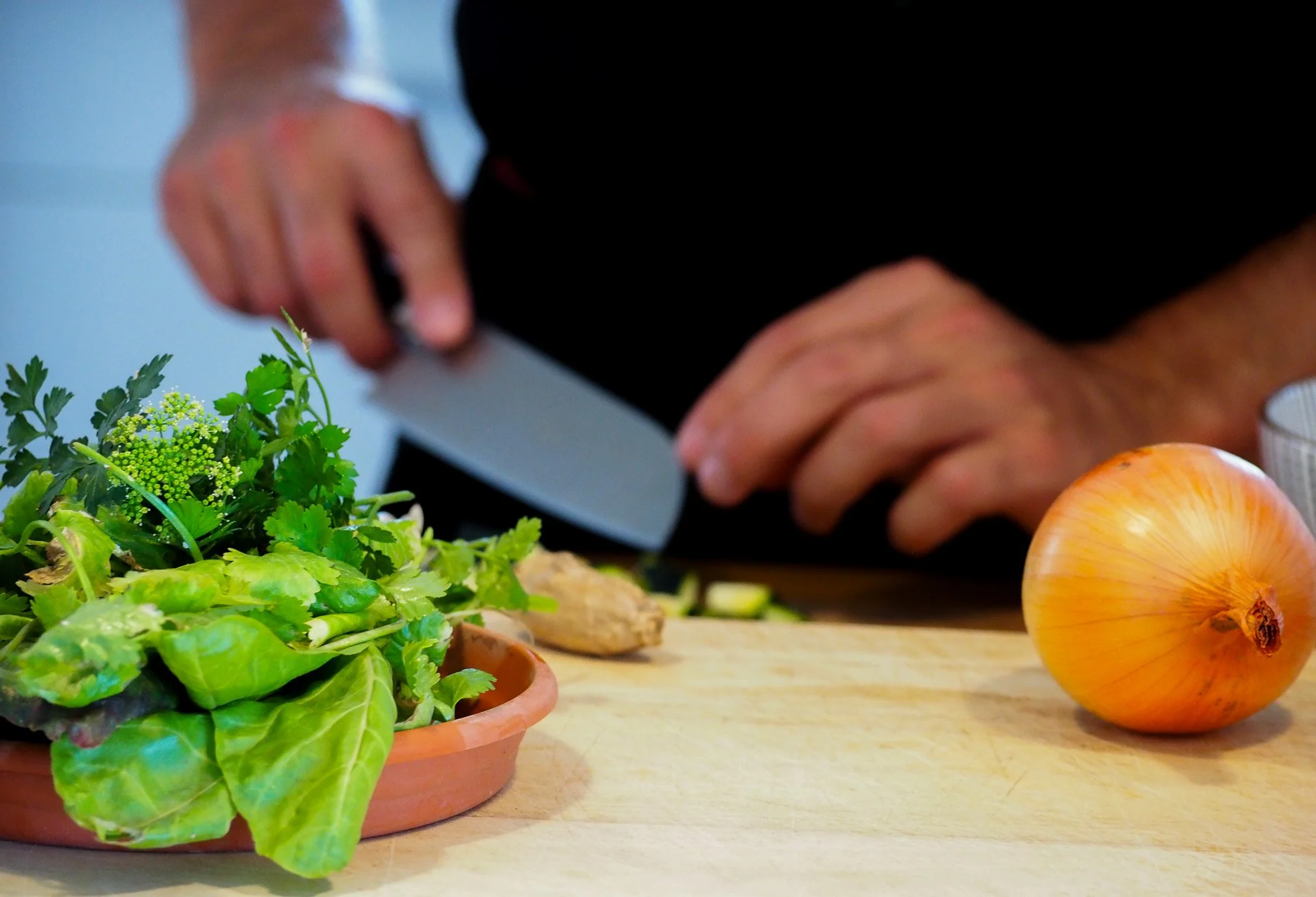 Person schneidet mit einem Messer auf einer Küchenarbeitsplatte. Vorne links befindet sich eine Topfpflanze mit grünen Blättern, rechts eine große Zwiebel, und im Hintergrund eine Hand, die mit einem Messer arbeitet.