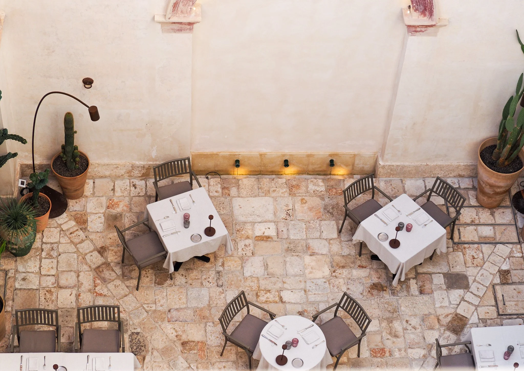 Innenaufnahme eines Restaurants mit mehreren Tischen, weißen Tischdecken, Besteck, Gläsern und Kerzen, gepflanzten Kakteen in Terrakottatöpfen, Mauerwand mit kleiner Beleuchtung, Steinboden.