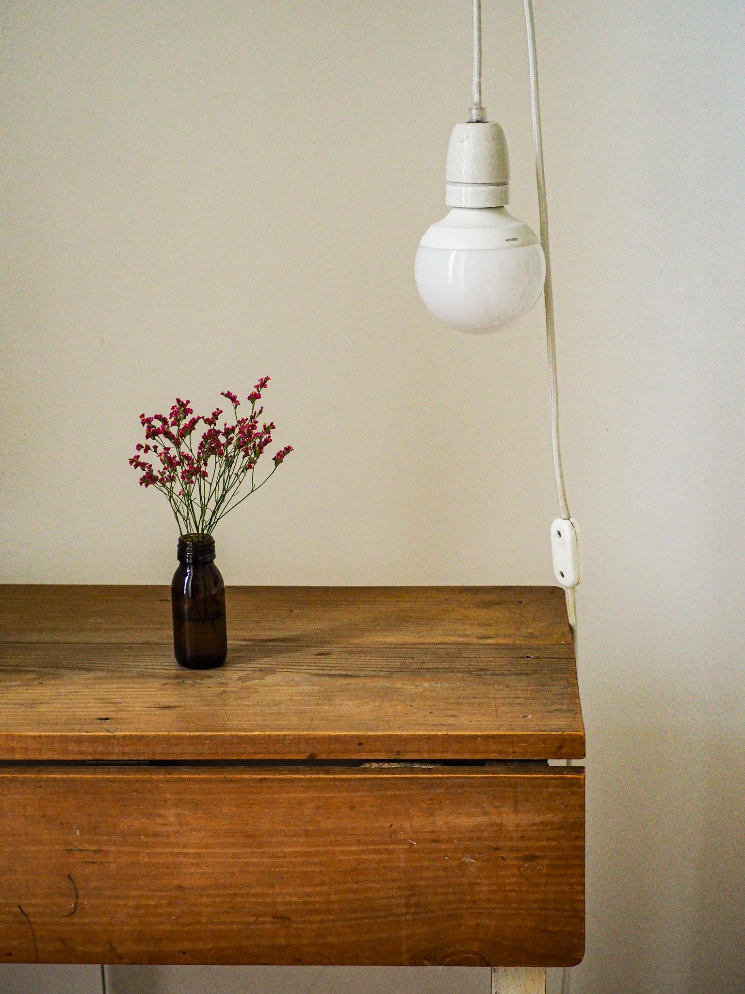 Ein braunes Glasflasche mit kleinen pinken Blumen steht auf einem Holztisch. An der Wand hängt ein weißer, gebauchter Glühbirnenlampen, die an einer weißen Steckdose angeschlossen ist.