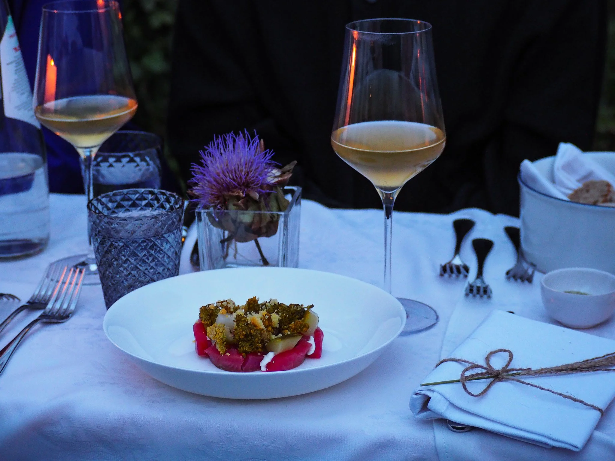 Tisch mit Wein, Wasser und einem Teller mit einem Gericht, Dekoration mit einer Blume, Besteck, ein weißer Stofftischläufer, gekreuzte Gabeln.