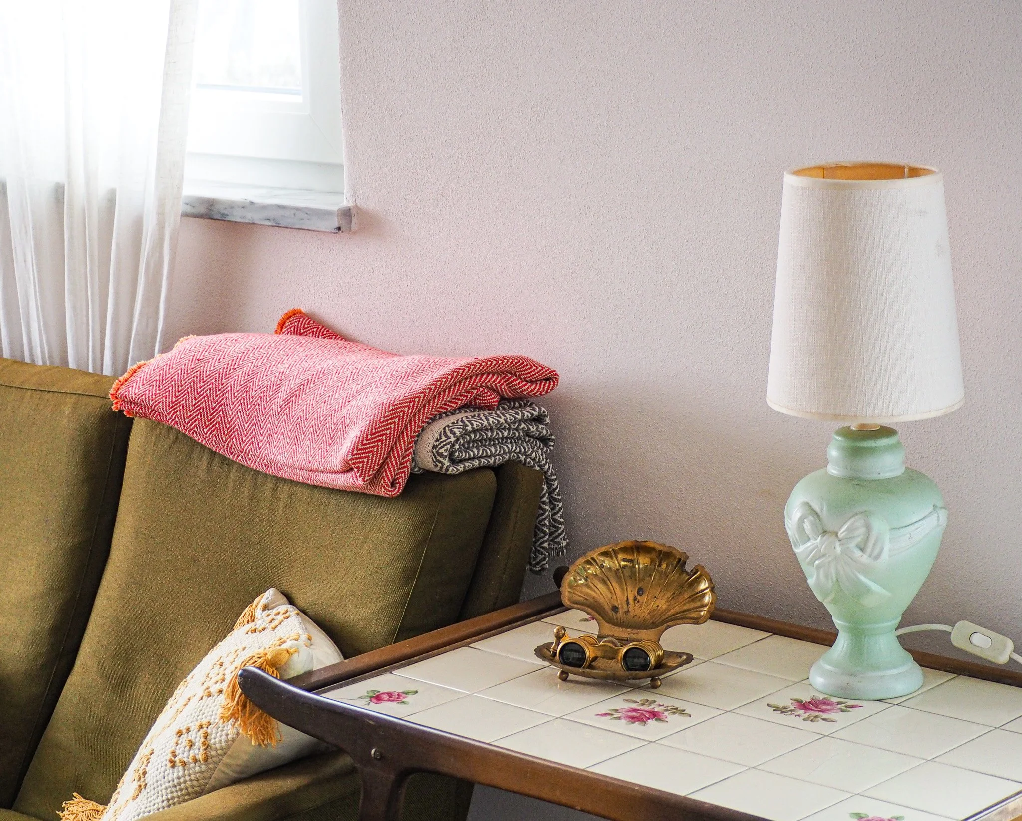 Wohnzimmer mit grüner Couch, Tisch mit Blumenmuster, weiße Lampe, Fenster mit weißen Gardinen, Decken auf der Couch, dekorative Gegenstände auf dem Tisch.