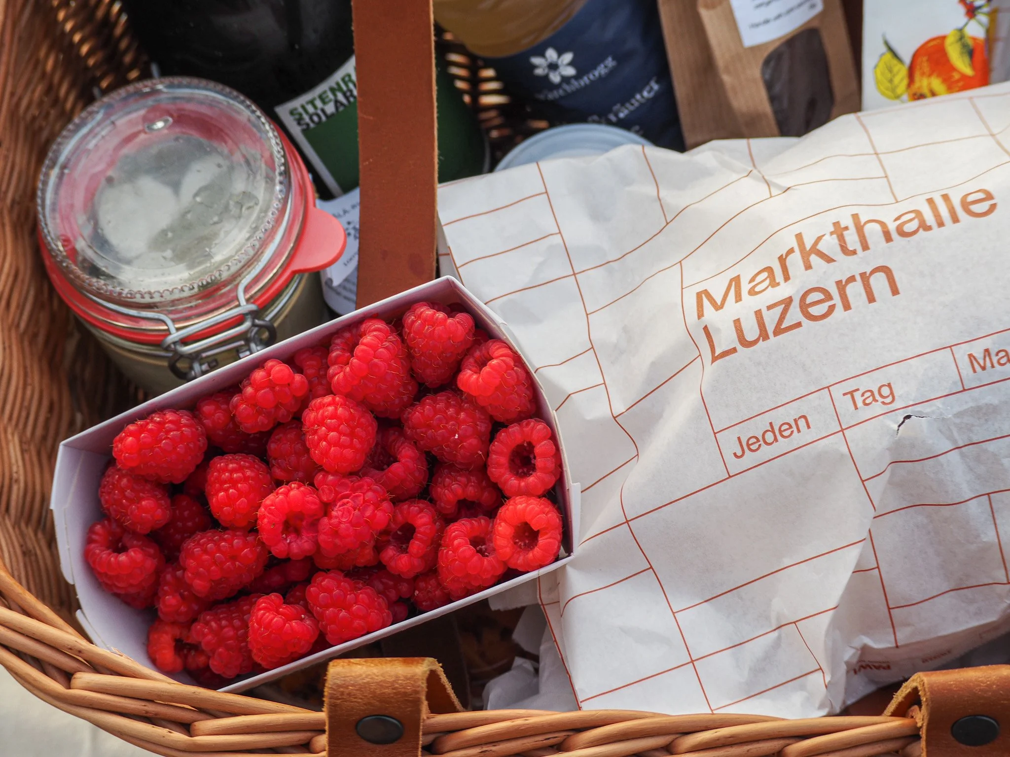Eine Einkaufstasche mit frischen Himbeeren, eine Glas Marmelade, eine Brottüte mit der Aufschrift "Markthalle Luzern" und einige Konservendosen.