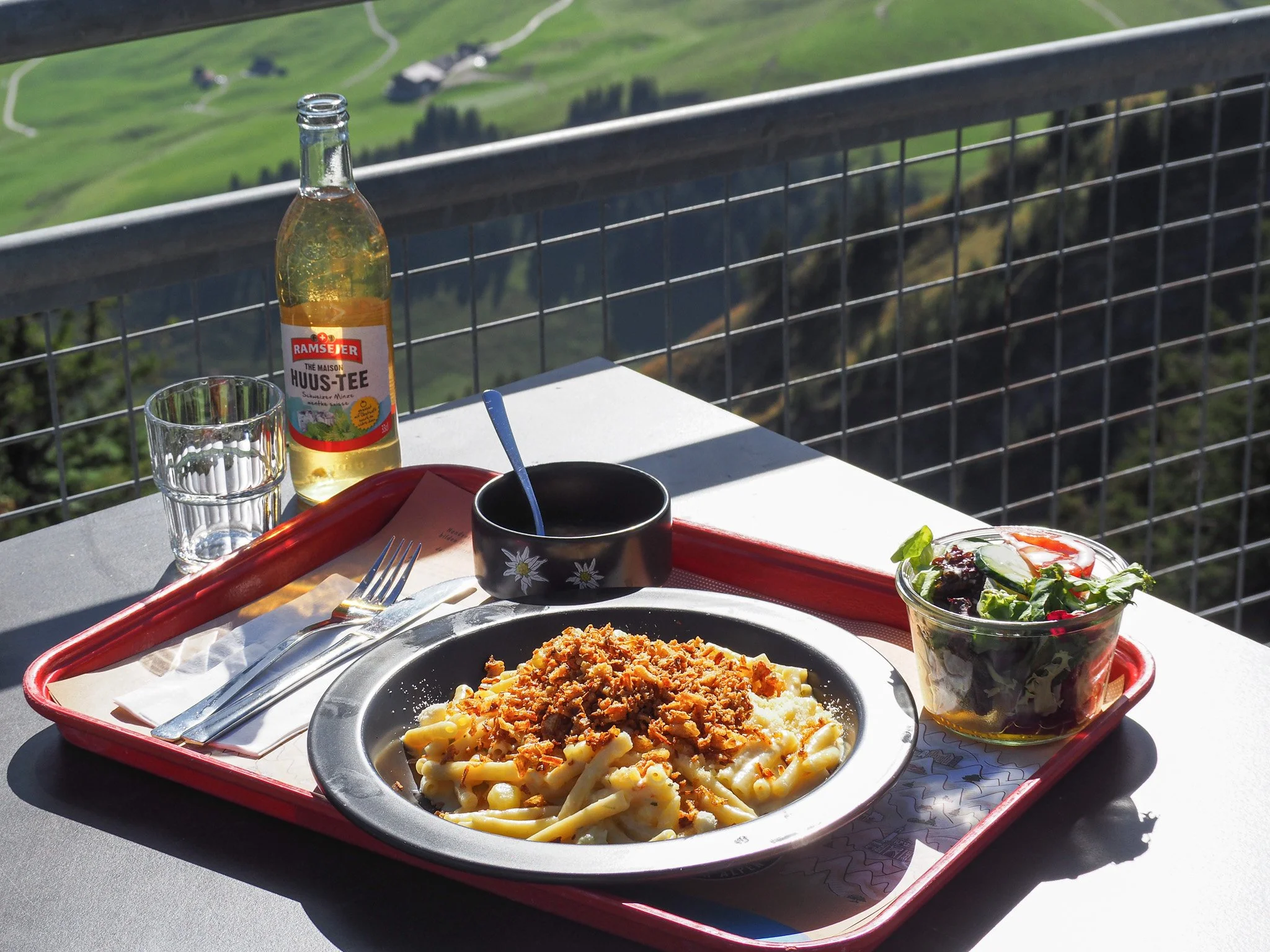 Mittagessen auf einem Teller mit Pasta und Hackfleisch, dazu ein grüner Salat in einer Glasschale, ein Glas Wasser, eine Flasche Tee und eine schwarze Tasse mit blumigem Muster auf einem roten Tablett auf einem Balkon mit Blick auf eine grüne Hügella