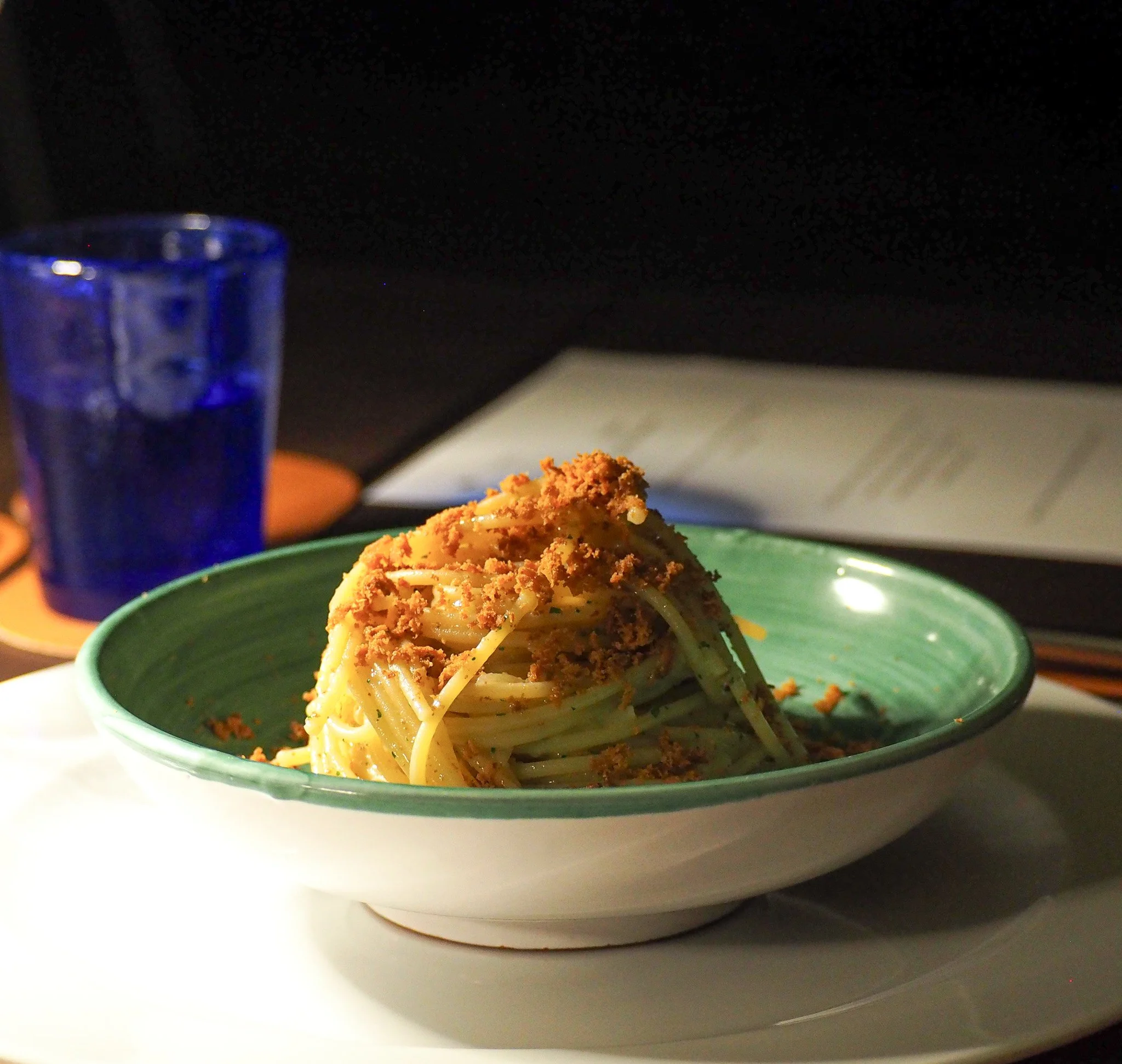 Nudeln in einer grünen Schüssel mit braunem Topping, auf einem weißen Teller, im Hintergrund ein blauer Wasserglas, eine dunkel gestaltete Oberfläche und ein aufgeschlagenes Buch.