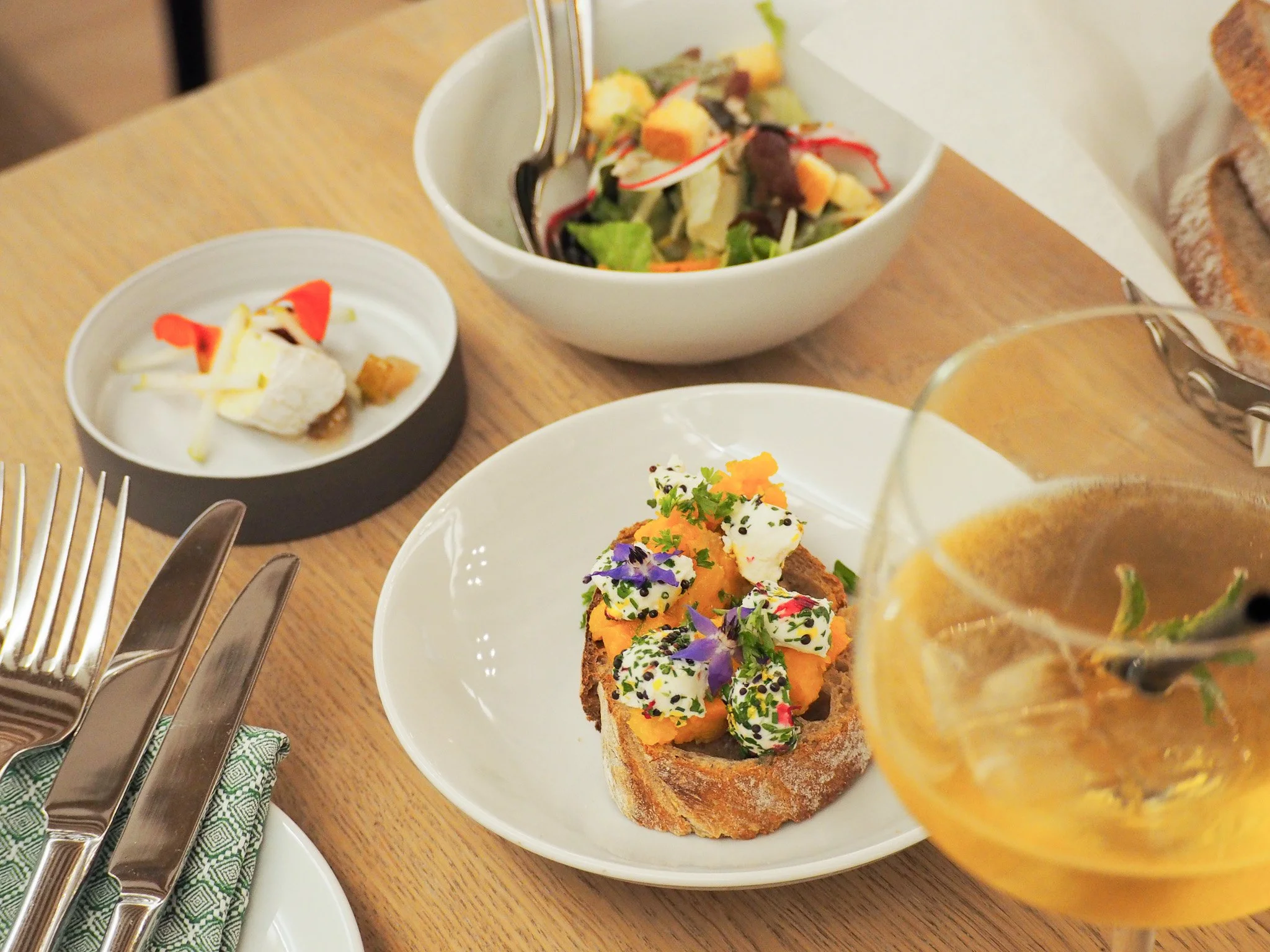 Ein gedeckter Tisch mit einem Teller mit belegtem Brot, einer Schüssel Salat, einer kleinen Schale mit Käse und Wurst, und einem Glas mit einem Getränk. Es gibt auch Besteck und ein Servietten auf dem Tisch.