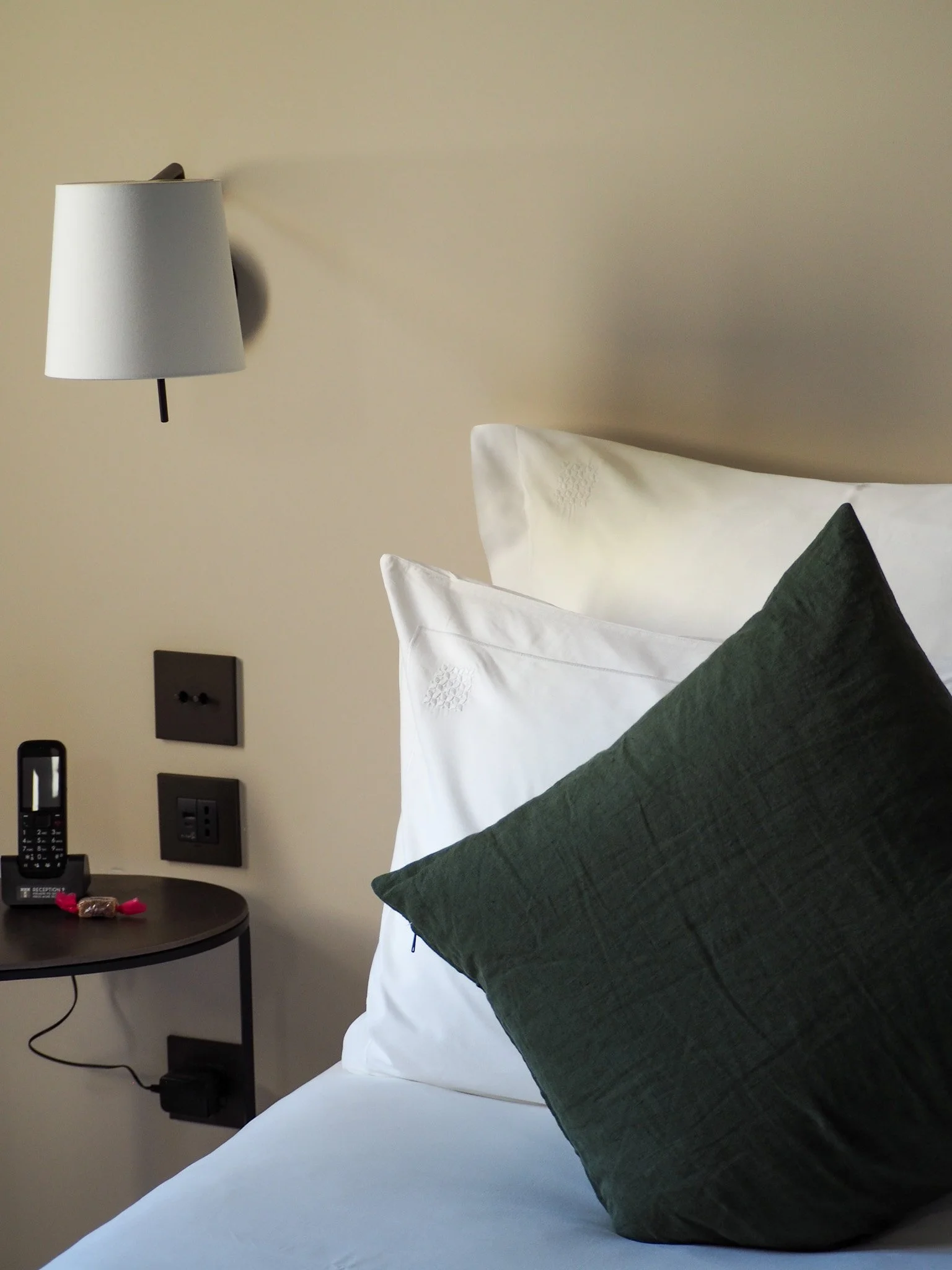 Ein Bett mit weißen Kissen und einem dunkelgrünen Kissen, ein Nachtisch mit einem Telefon und Schokolade, eine Wandleuchte an der Wand.