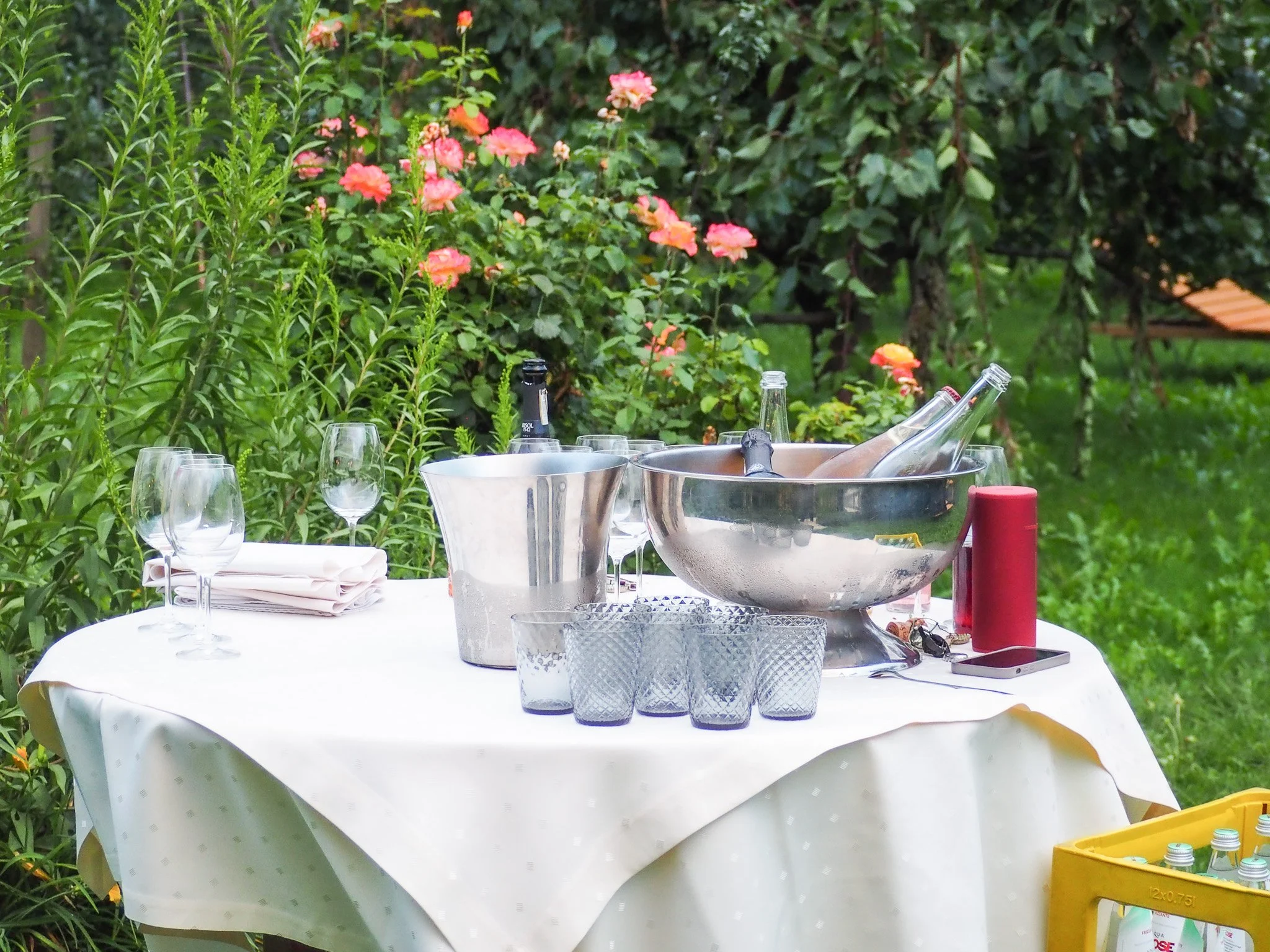 Ein Gartentisch mit weißen Tuch, Gläser, Flaschen, Eiskübel, Handy und Kerze steht im Garten, umgeben von Blumen und Bäumen.