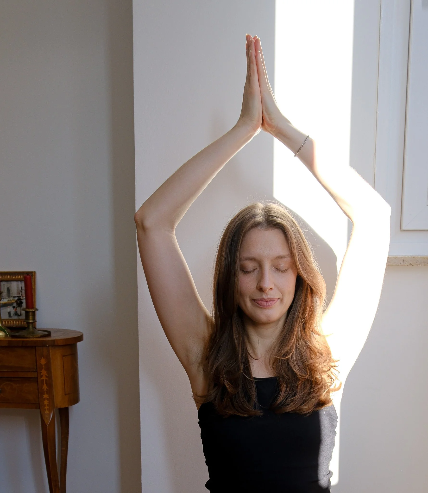 Eine junge Frau macht Yoga im hellen Raum, die Hände sind in der Gebetshaltung über ihrem Kopf, Augen geschlossen, entspannter Gesichtsausdruck.