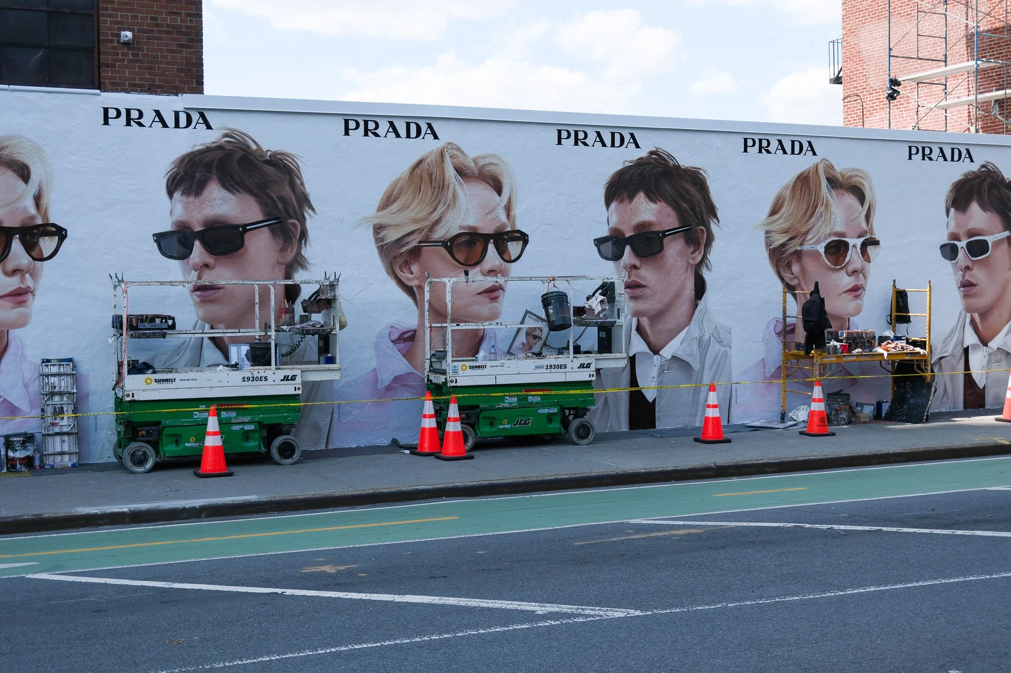 Großwand mit einem Werbeplakat für Prada, das mehrere Frauen mit unterschiedlichen Sonnenbrillen zeigt, während auf der Straße darunter Baugeräte und Verkehrsleitkegel sind.