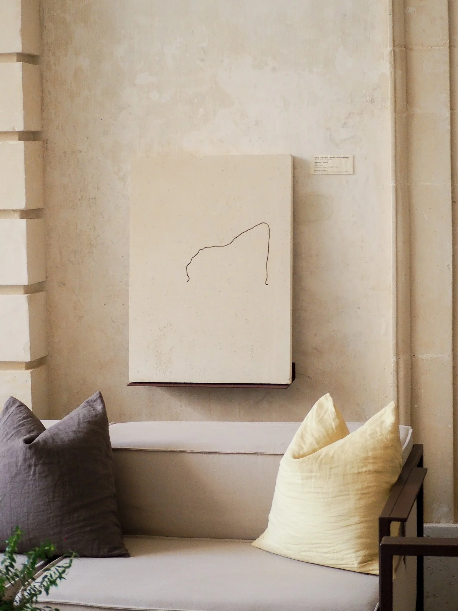 Einichel weißer Sitzbereich mit grauen und cremefarbenen Kissen, gegenüber eine weiße Wand mit abstraktem Kunstwerk mit einer schwarzen Linie. Es sieht aus wie ein minimalistisches Kunstwerk, das an einem ruhigen und eleganten Raum hängt.